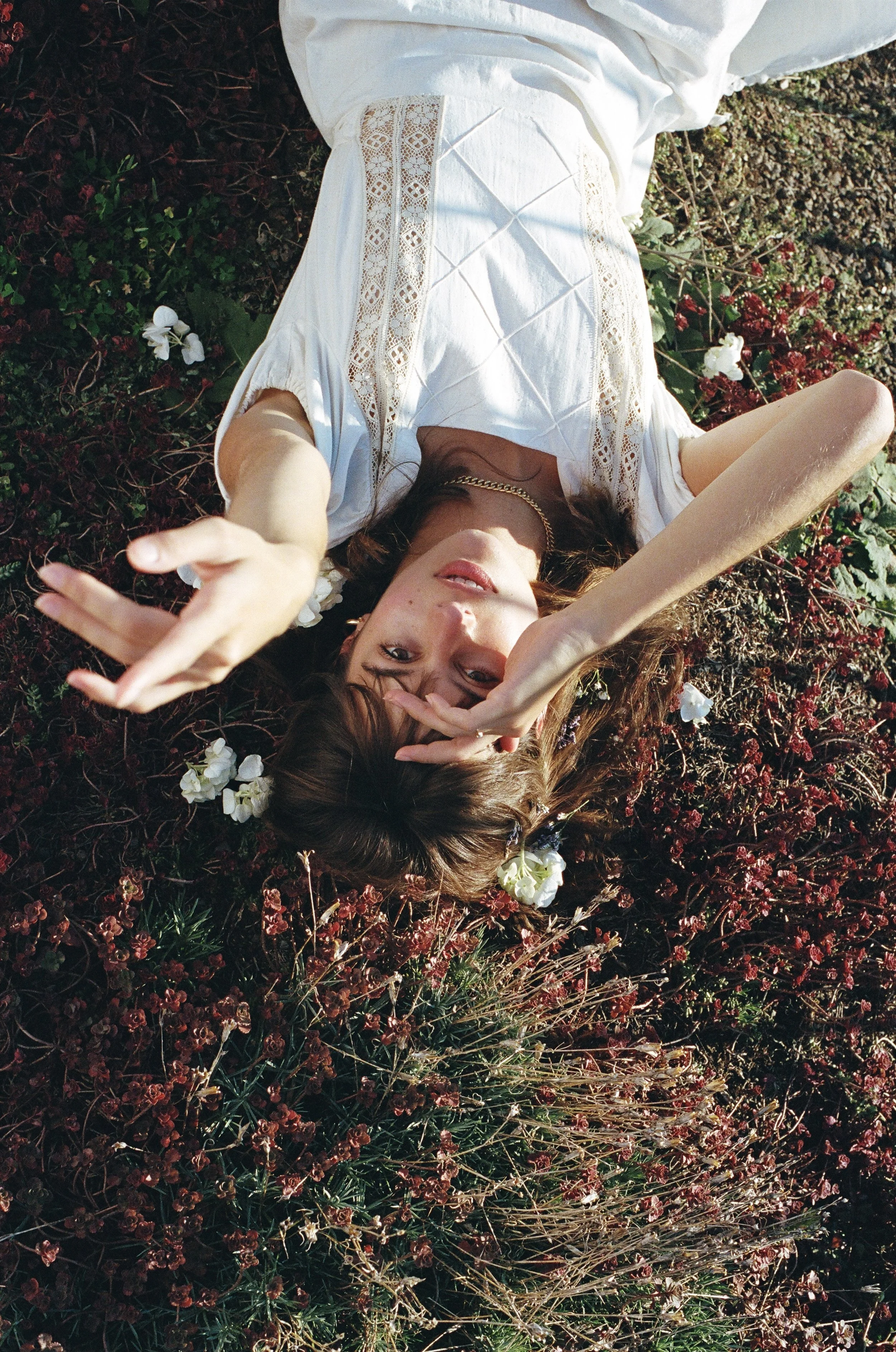 A young woman with brown hair and light skin lies on her back amidst red and green plants, wearing a white dress with lace details, making a peace gesture with one hand near her face and the other extending outward.