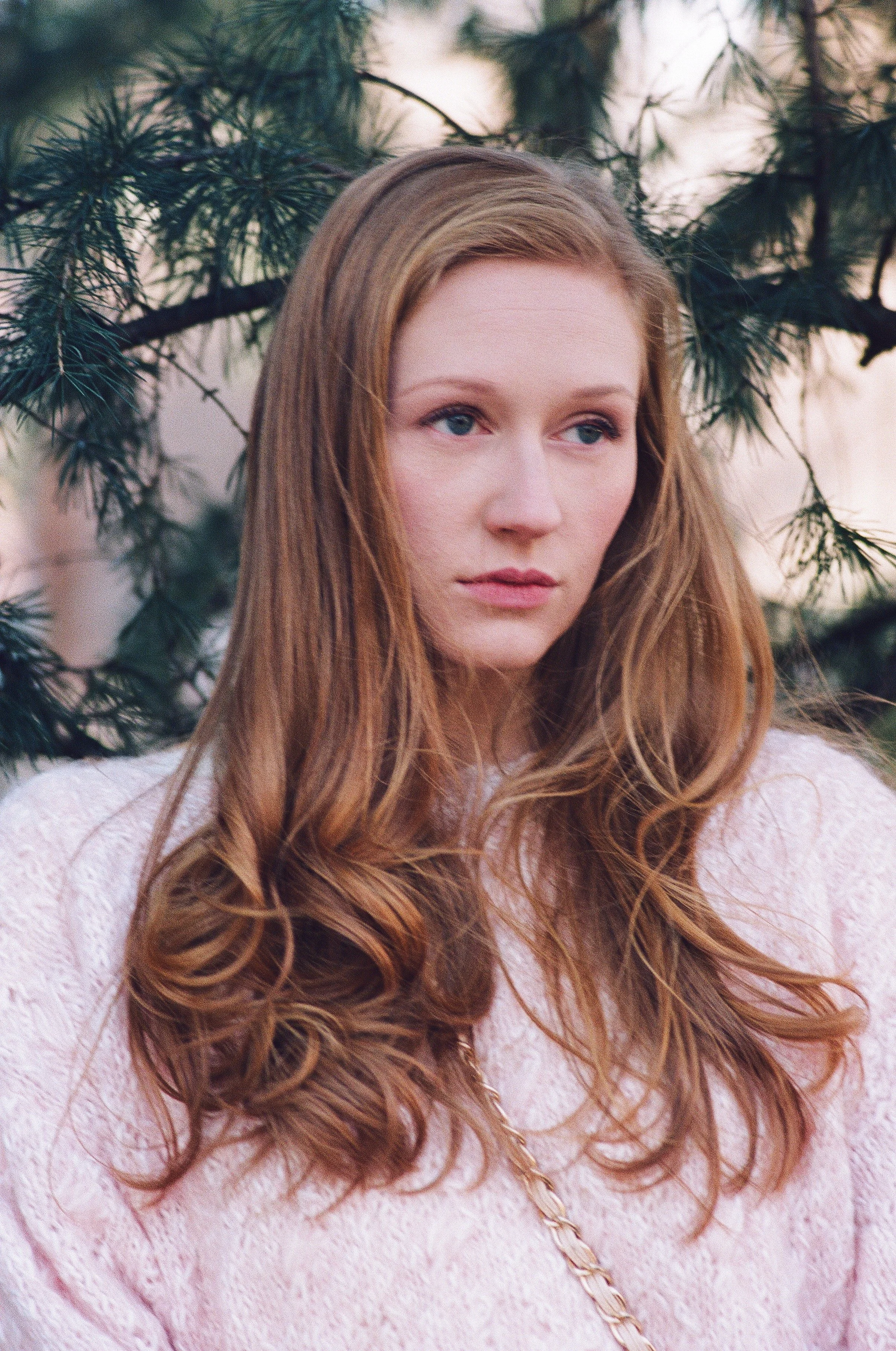 A woman with long, wavy red hair and light skin standing outdoors near green pine tree branches.