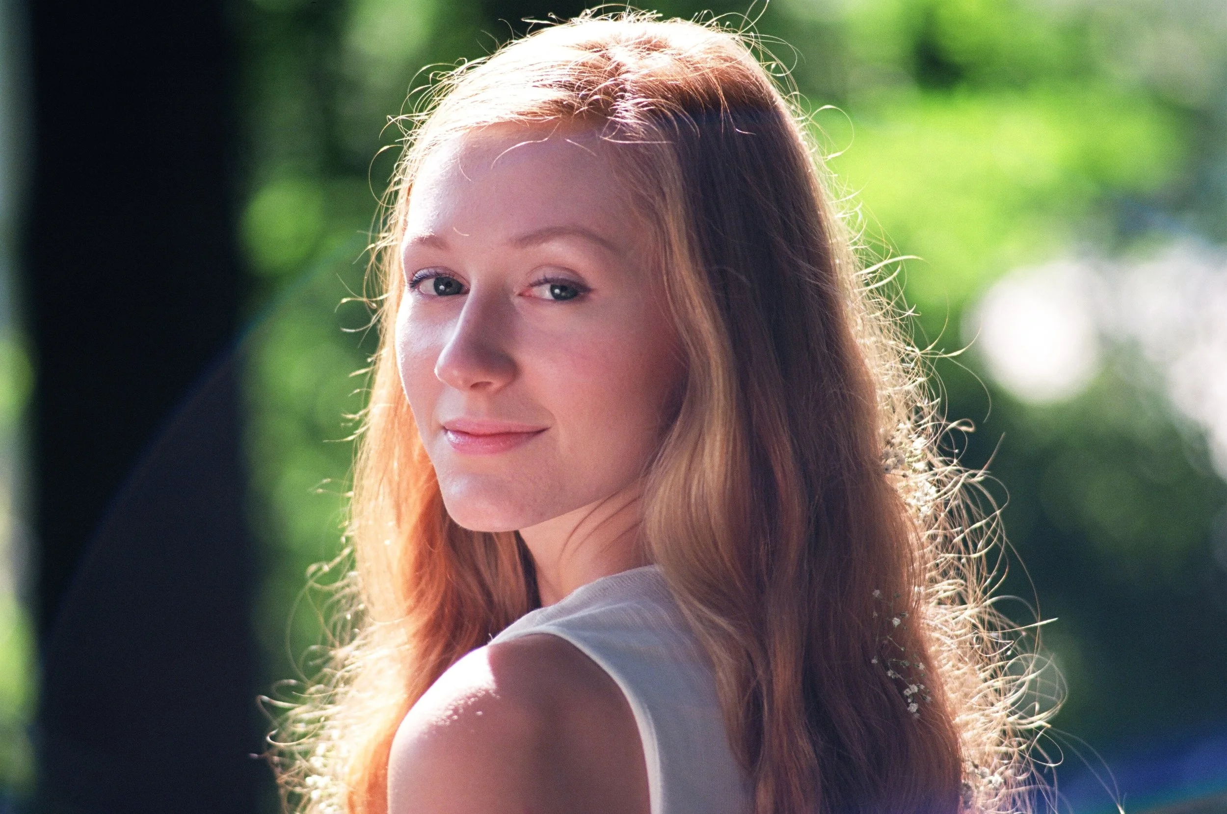 A young woman with long wavy red hair and fair skin stands outdoors on a sunny day, with blurred green foliage in the background, smiling softly at the camera.