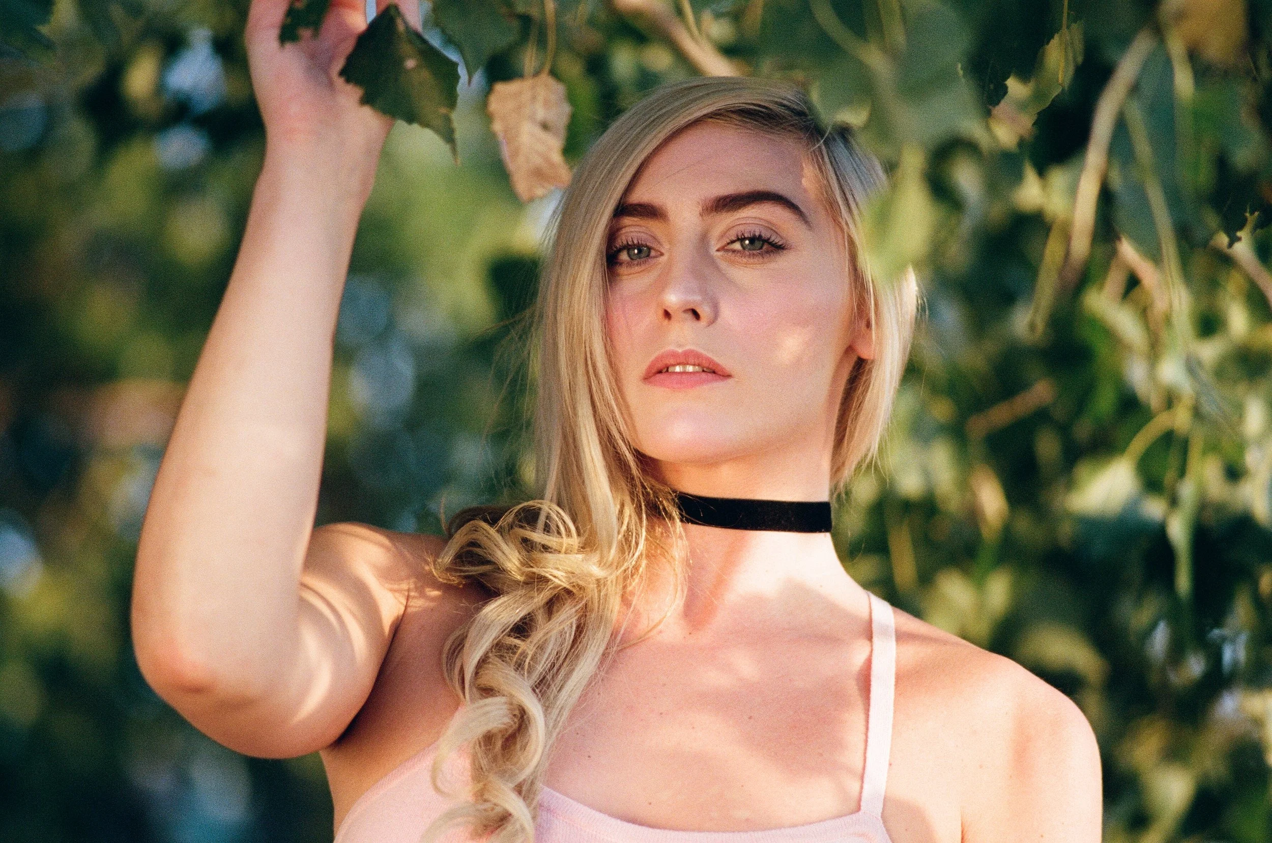 A young woman with blonde hair, wearing a pink top and a black choker, standing outdoors among green leaves, looking into the camera.
