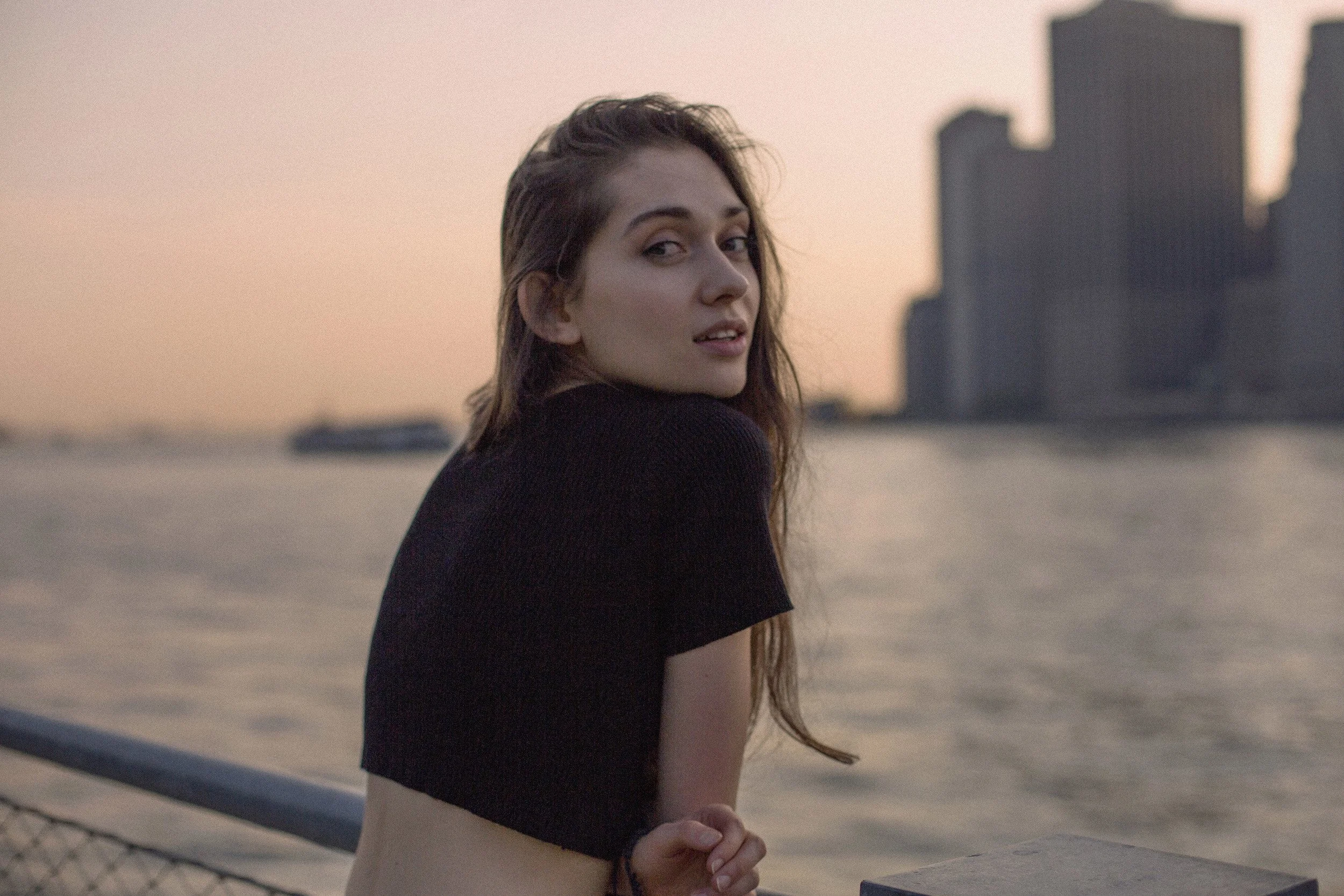 A young woman with long brown hair looking back over her shoulder, standing by the water with a city skyline in the background during sunset.