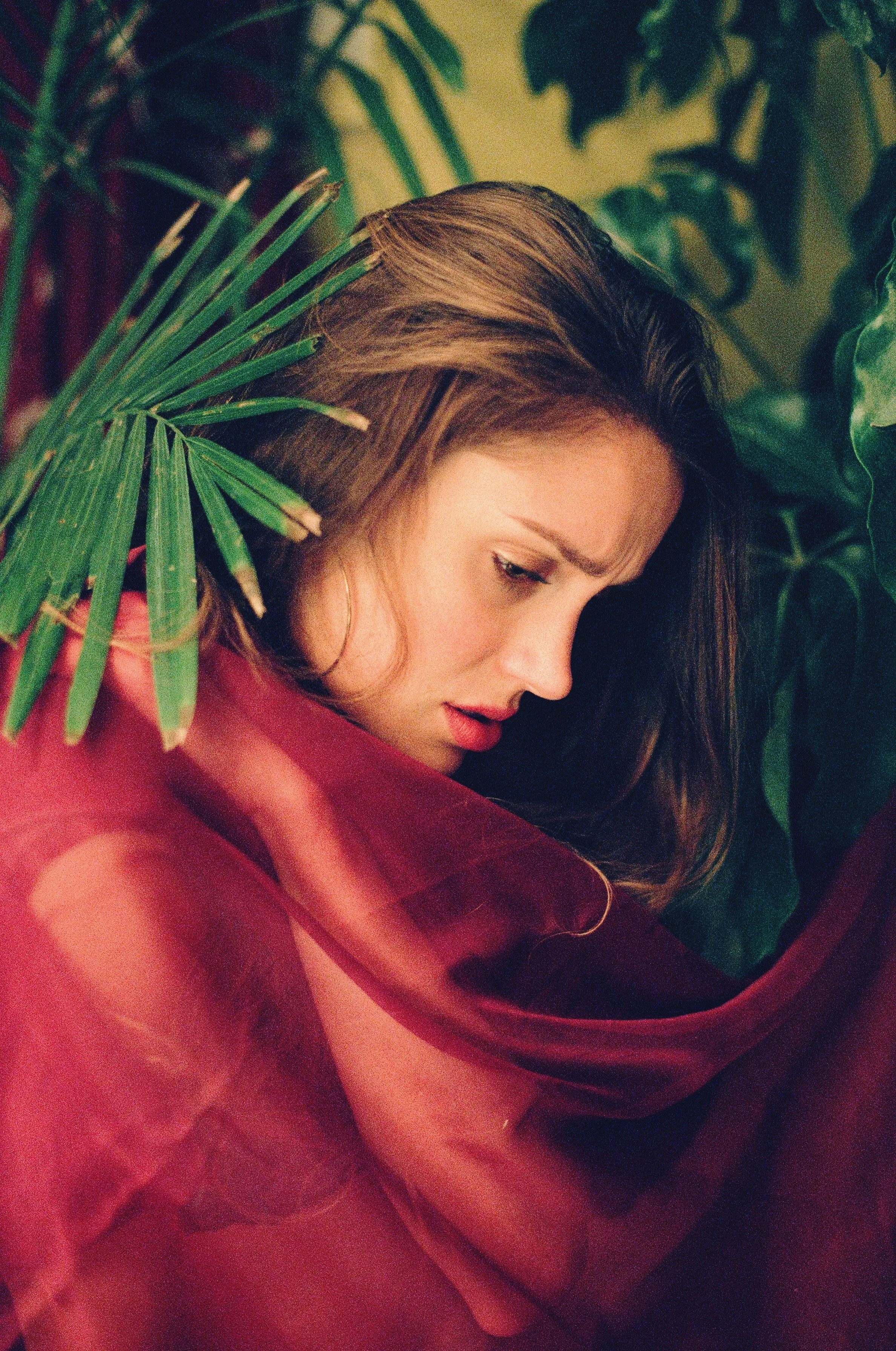 A woman with long brown hair and light makeup, looking down and to the side, surrounded by green tropical leaves and draped in a sheer, red fabric.
