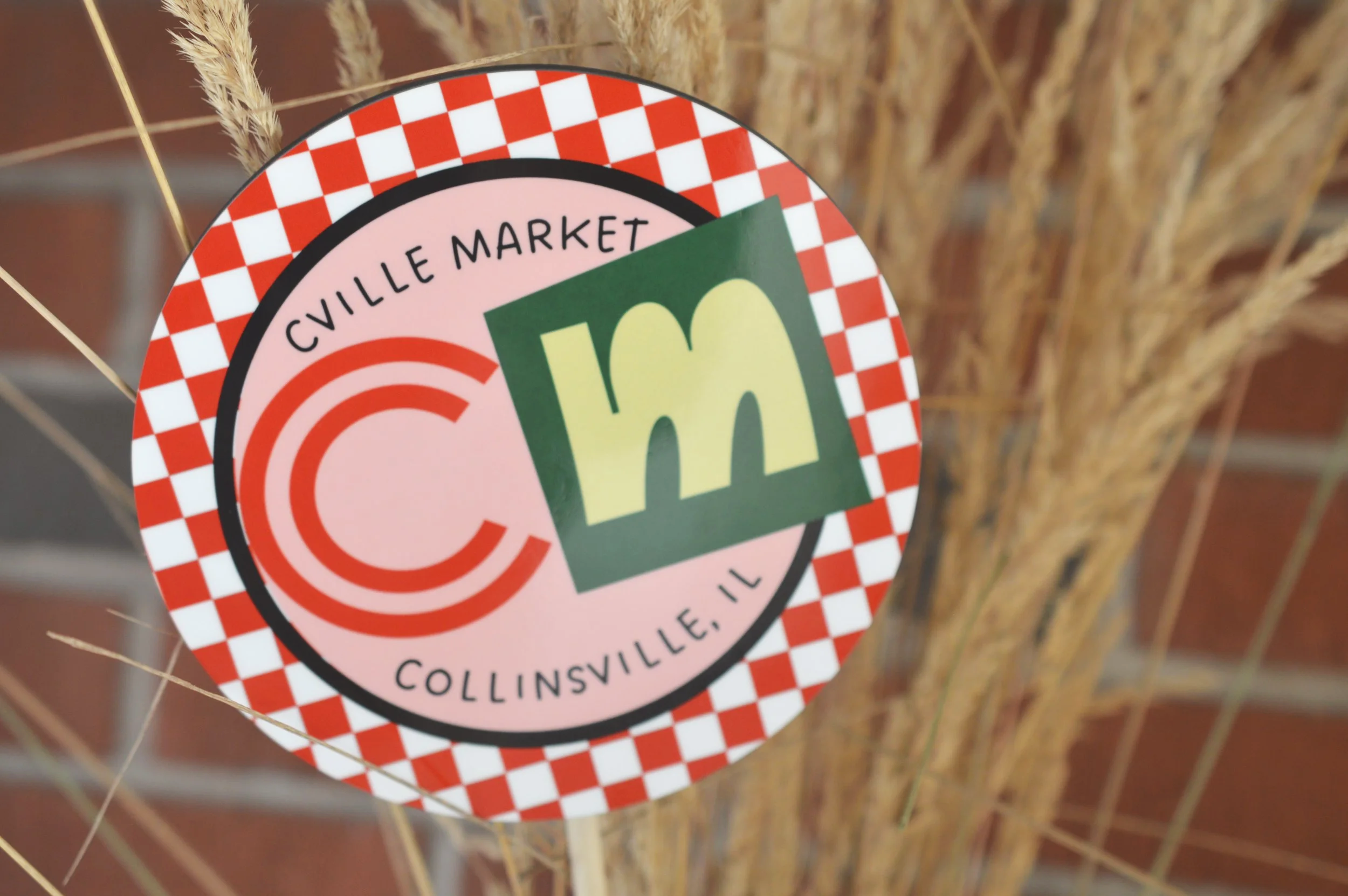 Close-up of a circular sticker with a red and white checkered border, featuring the text 'Cville Market' and 'Collinsville, IL' with a logo in the center that includes a green square with a stylized yellow 'm' and a red concentric circle design.