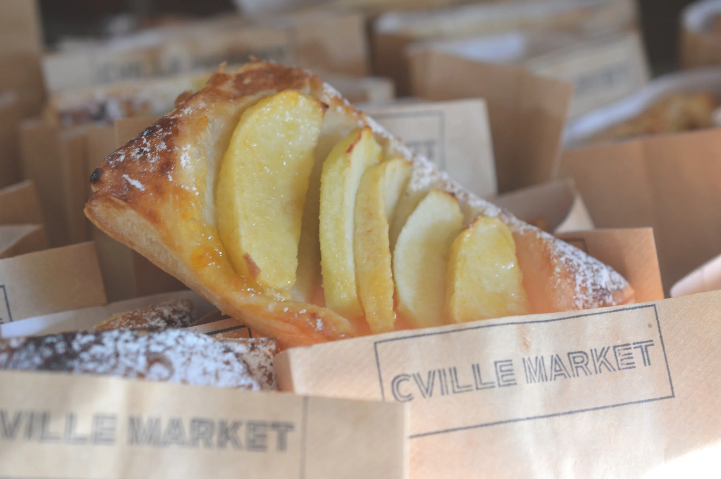 A piece of apple tart with sliced apples inside, dusted with powdered sugar, presented in a paper wrapper labeled 'VILLE MARKET'.
