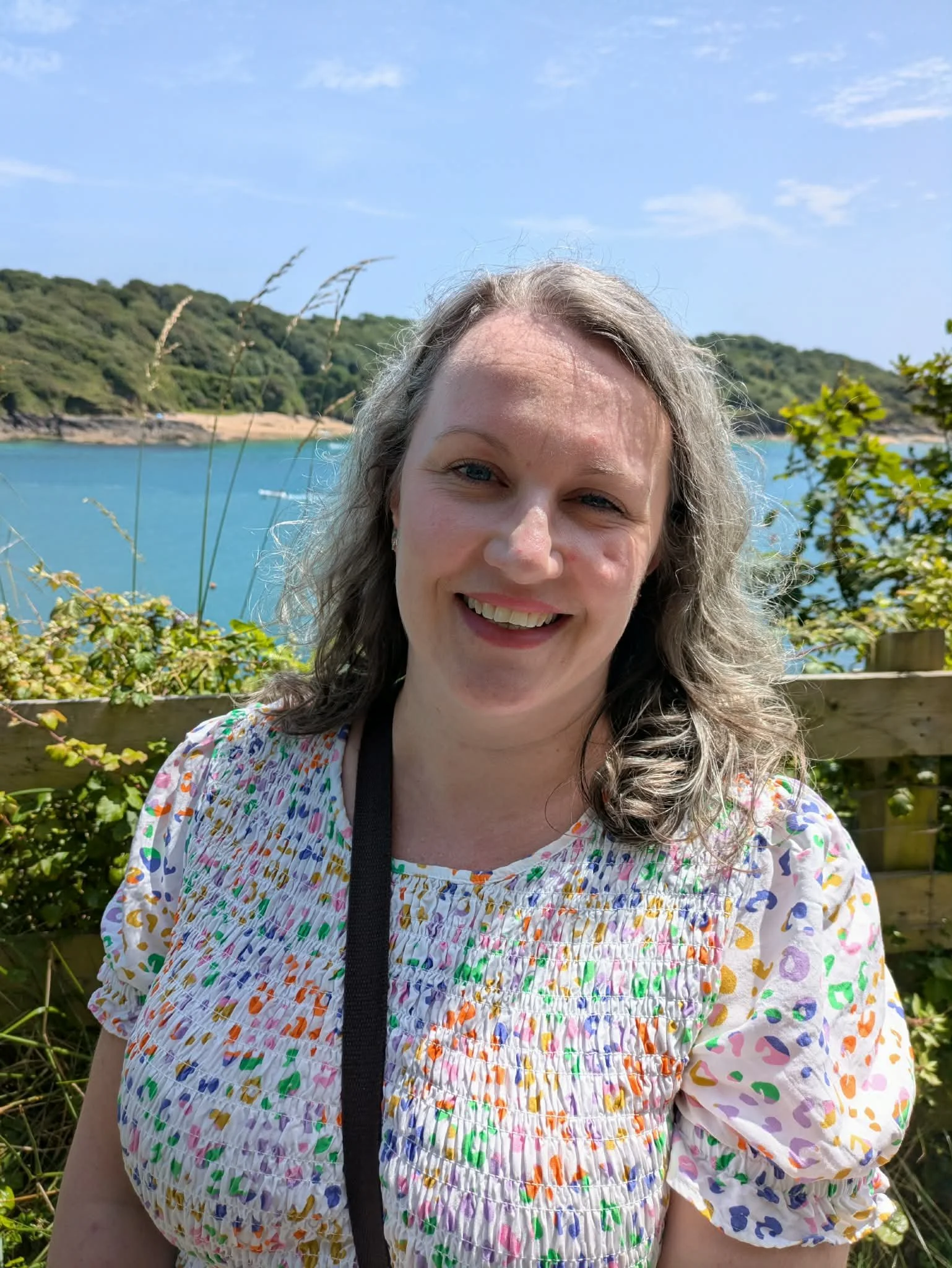 A woman with curly gray hair smiling outdoors near a body of water, with a wooded hill in the background, wearing a colorful, floral-patterned blouse.