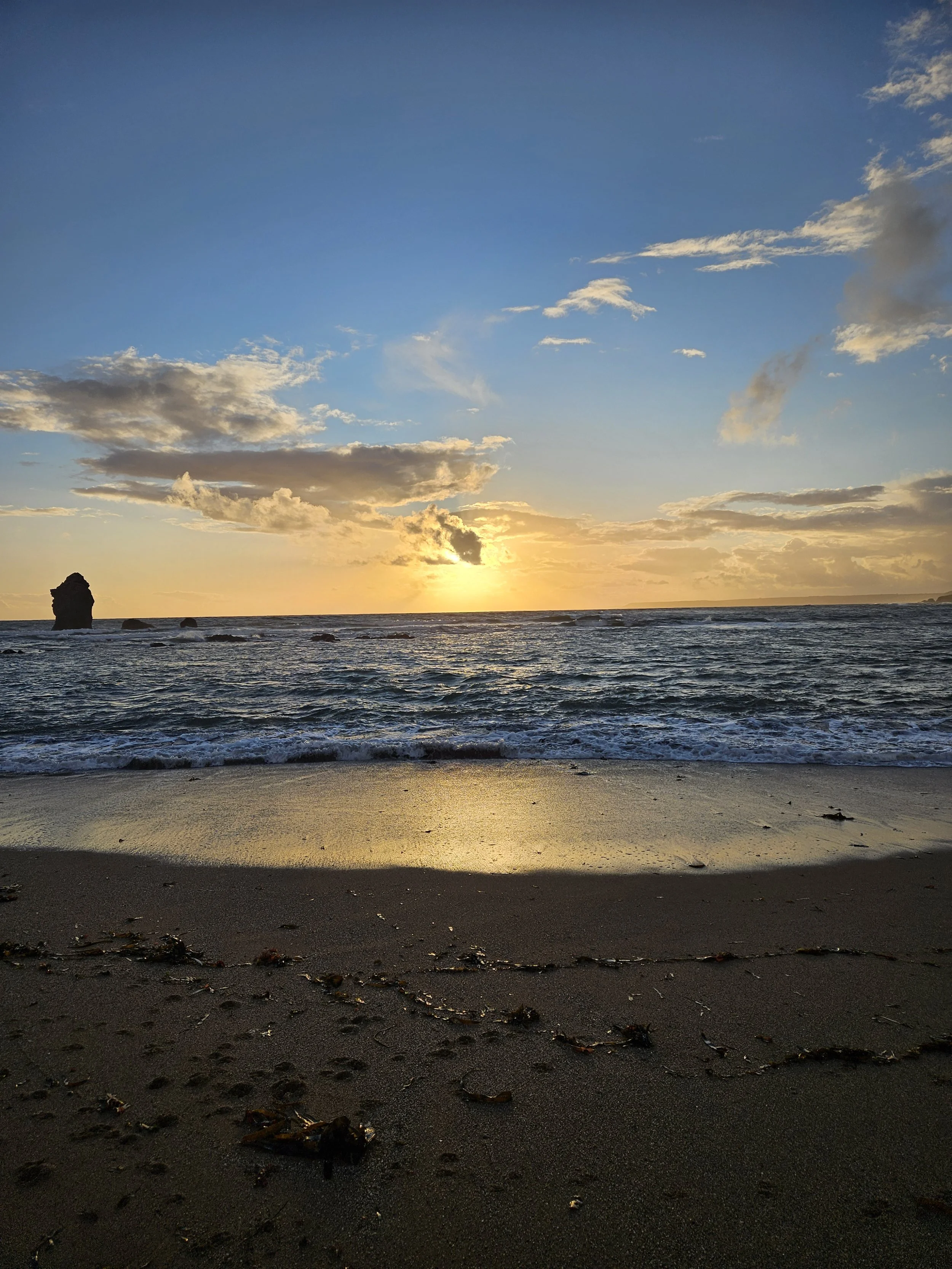 Sunset over the ocean with some clouds in the sky, rocks in the water, and seaweed on the sandy beach