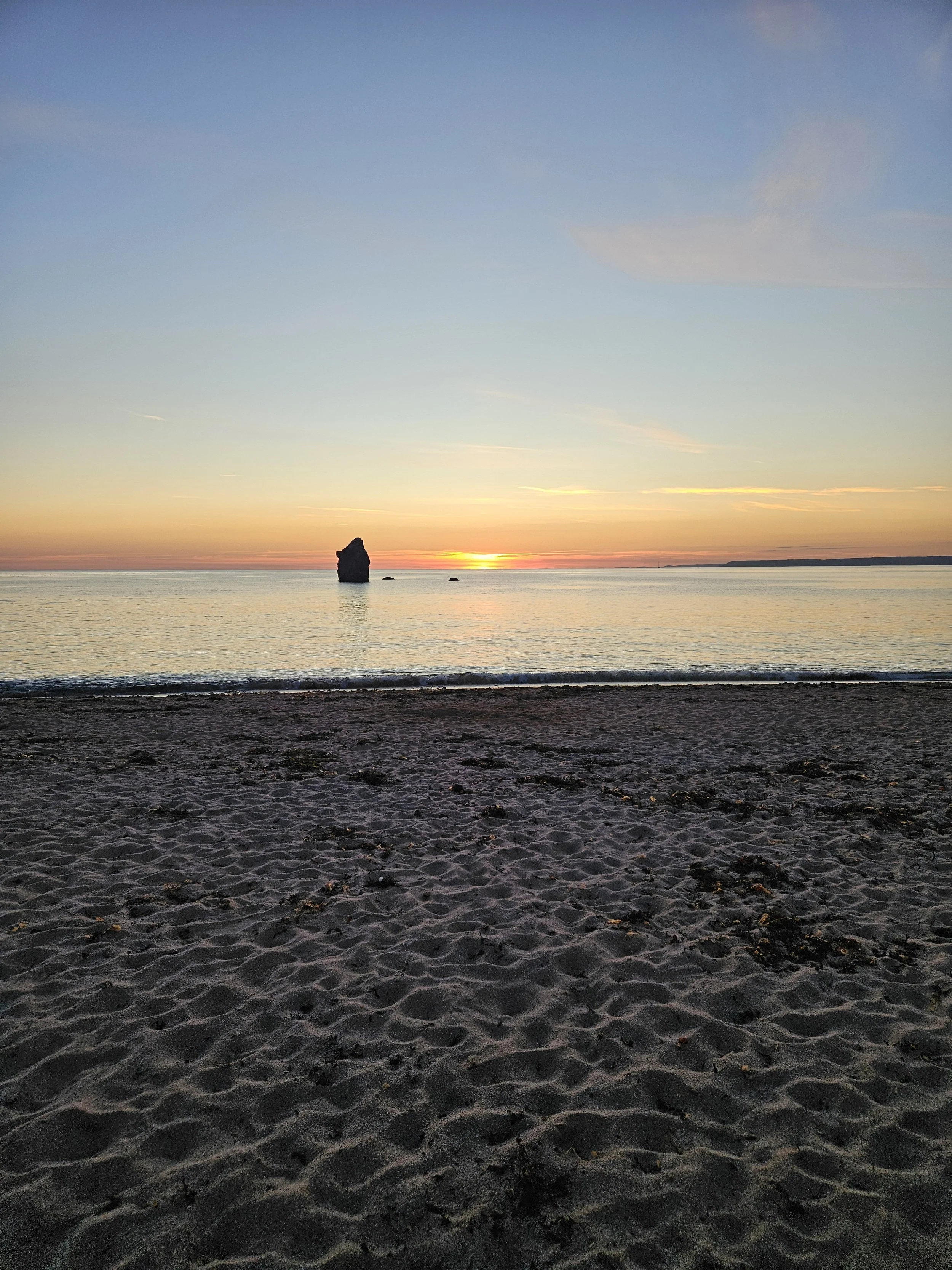 A beach at sunset with a large rock formation in the water and a distant landmass on the horizon. The sky has soft clouds with pastel colors from the setting sun.