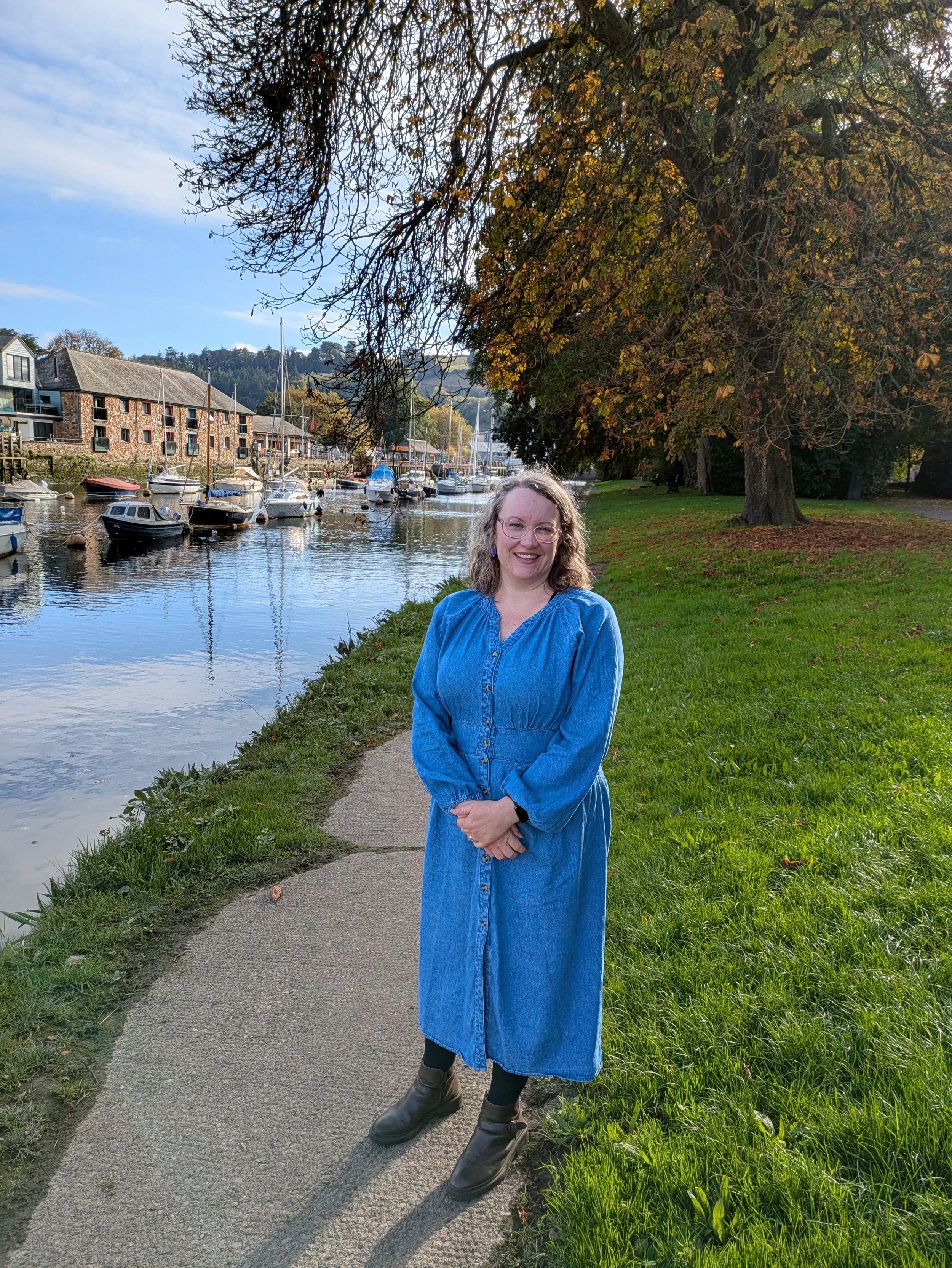 A woman standing on a sidewalk near a river with boats docked along the water, under a large tree with autumn leaves, smiling at the camera.