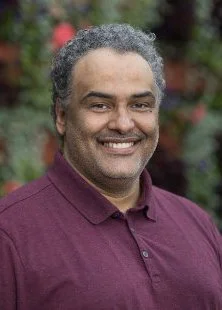 A smiling man with gray curly hair wearing a maroon polo shirt outdoors, with blurred greenery in the background.