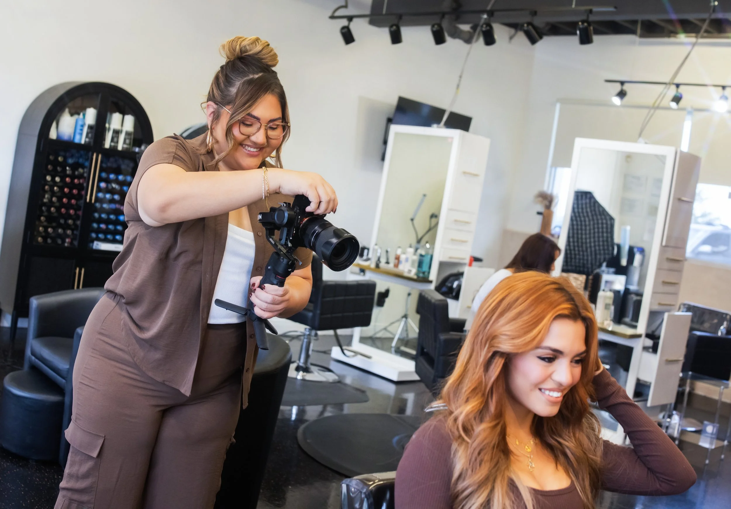 A hairstylist with glasses and a bun is trimming a woman's long, wavy, red hair in a modern salon. The woman is smiling, dressed in a brown top, sitting in a salon chair. The background features hair products on shelves, mirrors, and salon equipment.