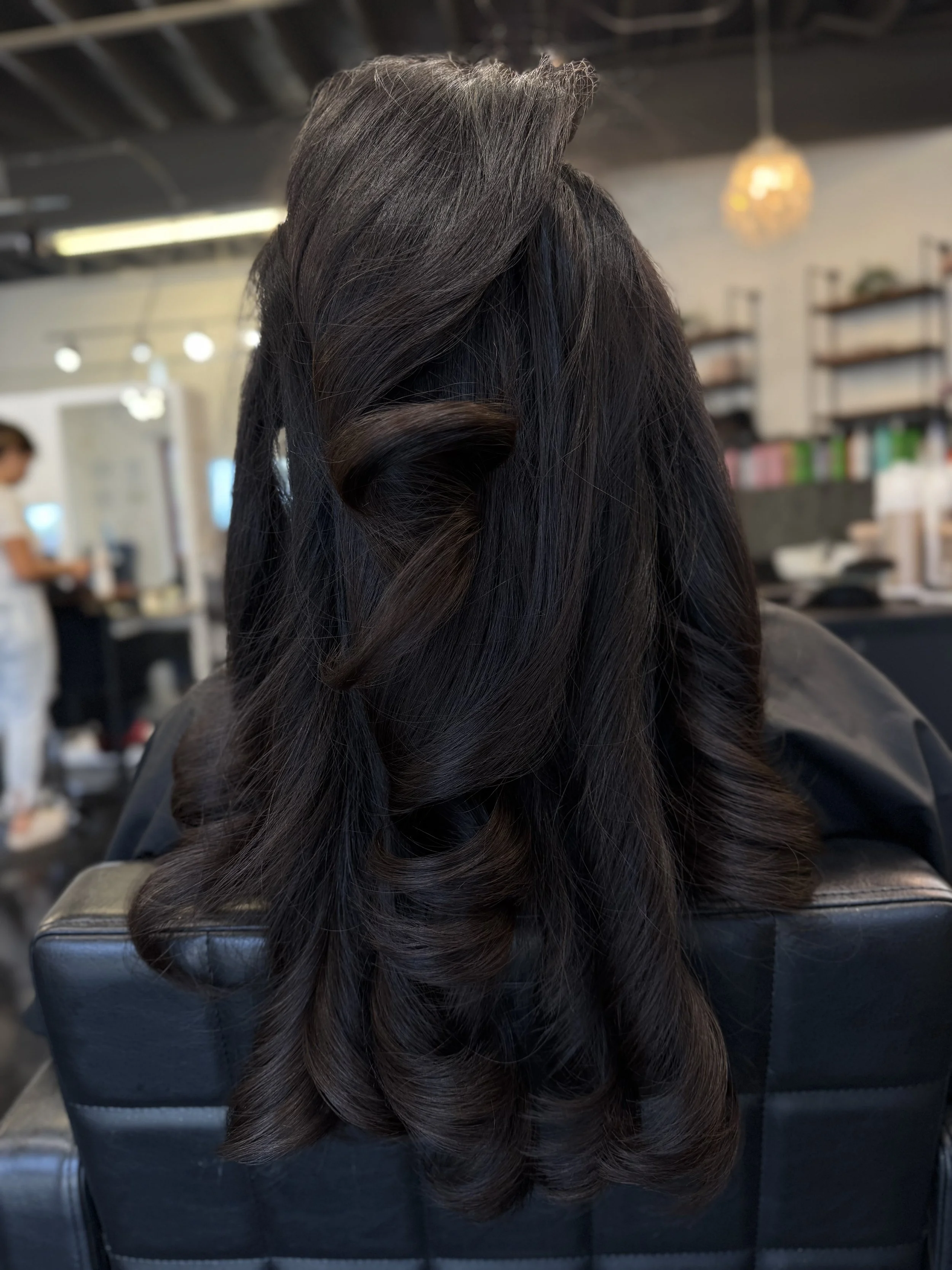 Well-styled long dark brown hair with soft curls, sitting on a black salon chair in a hair salon.