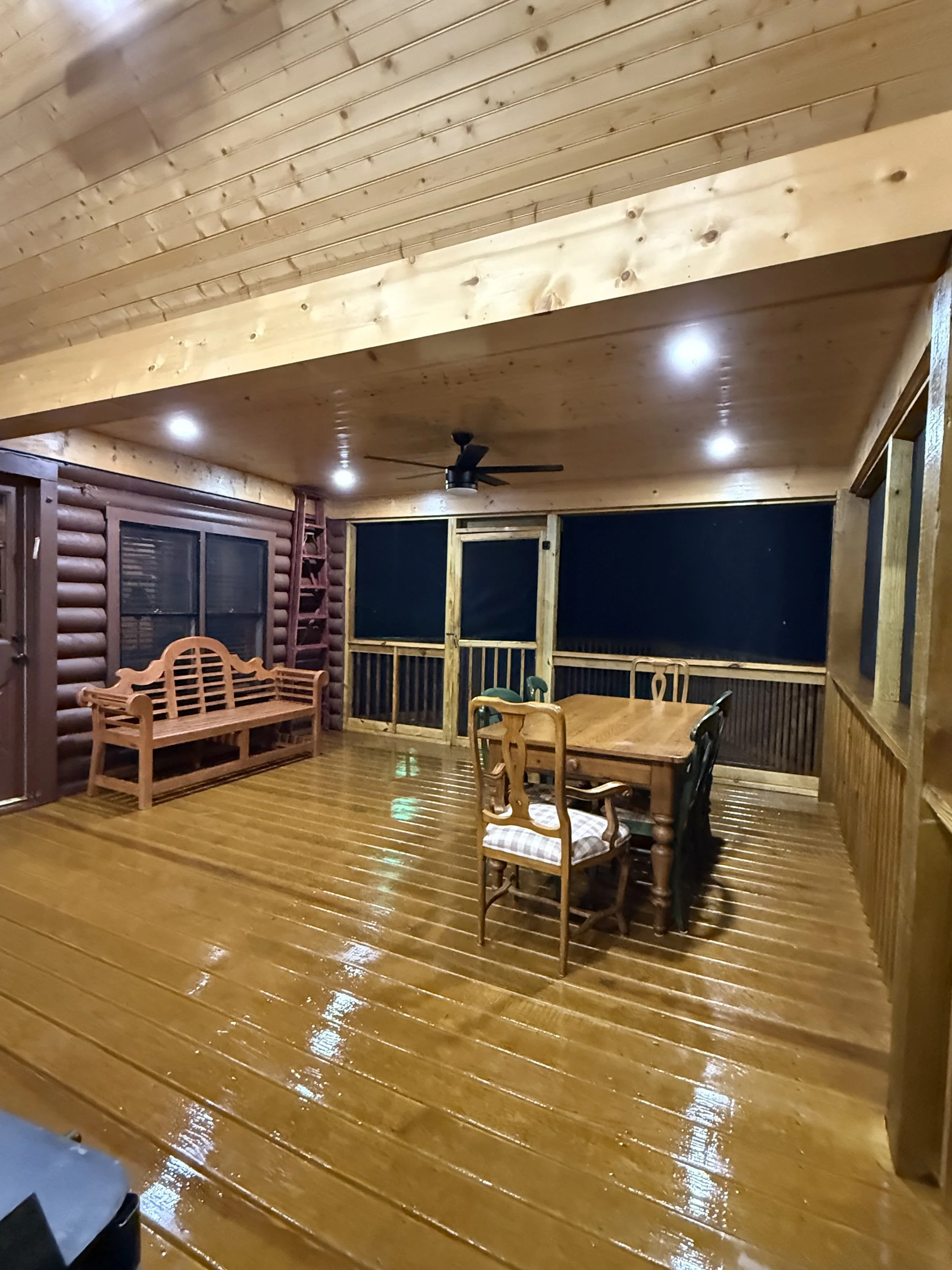 Interior of a wooden porch or deck with wooden flooring, ceiling, and walls, featuring a wooden bench, a dining table with chairs, and a ceiling fan.