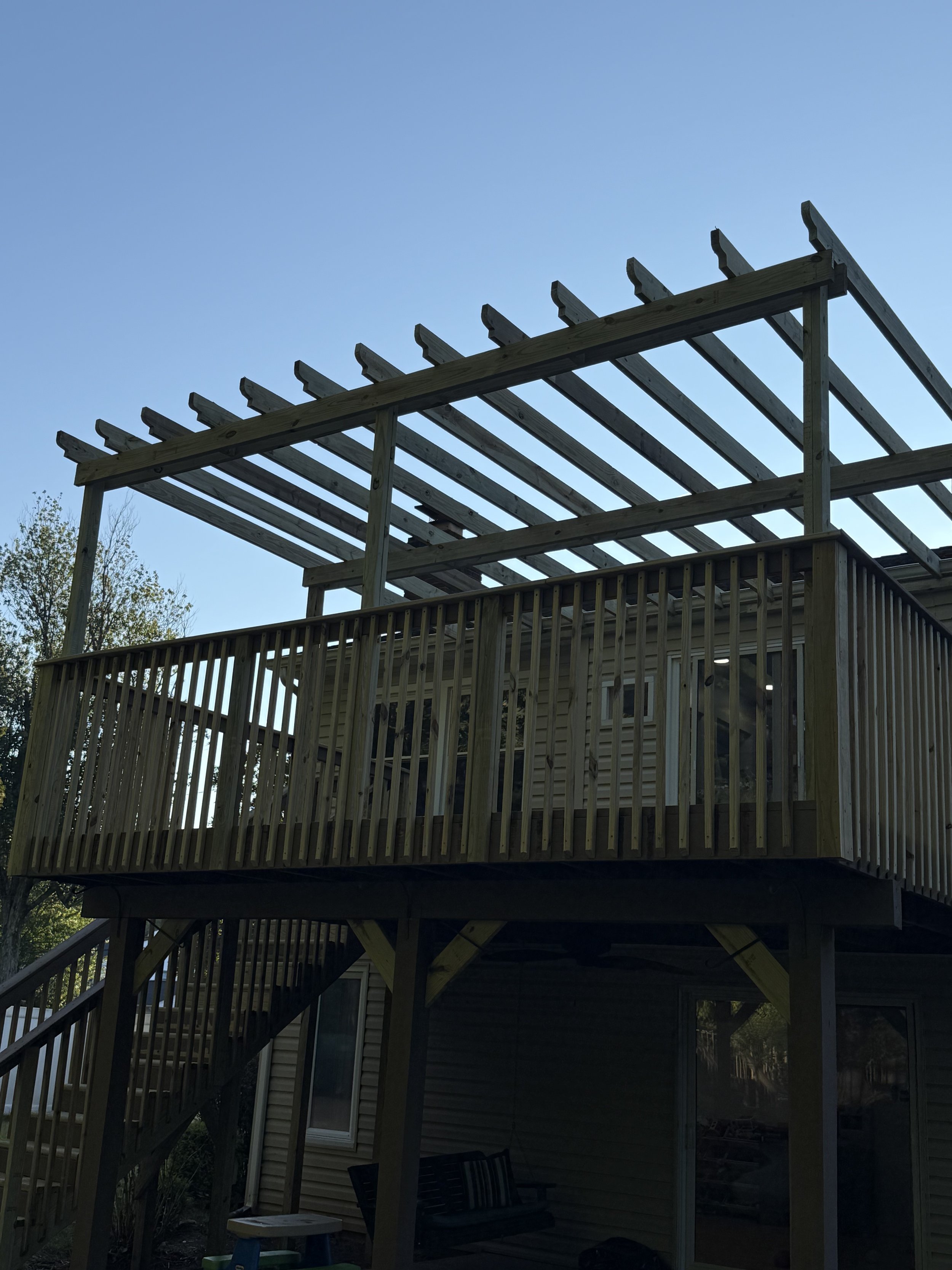 Wooden deck under construction with open roof framework, attached to a house with stairs leading up to it, set against a clear blue sky.