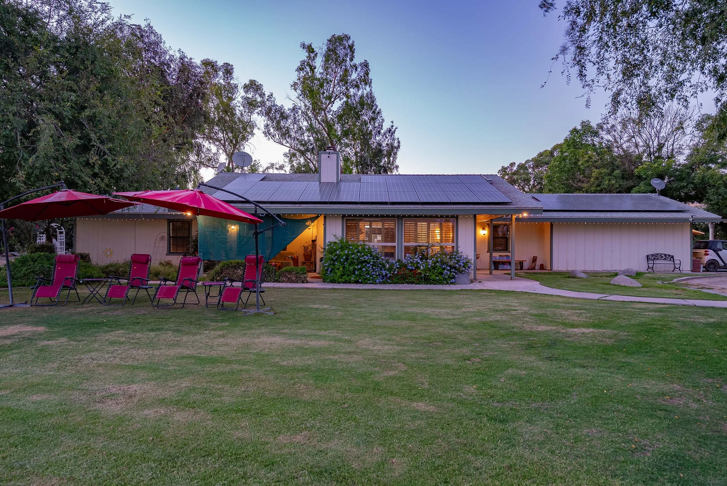 A single-story house with solar panels on the roof, illuminated from the inside, surrounded by a well-maintained lawn with outdoor seating, umbrellas, and tables, set during dusk with trees in the background.