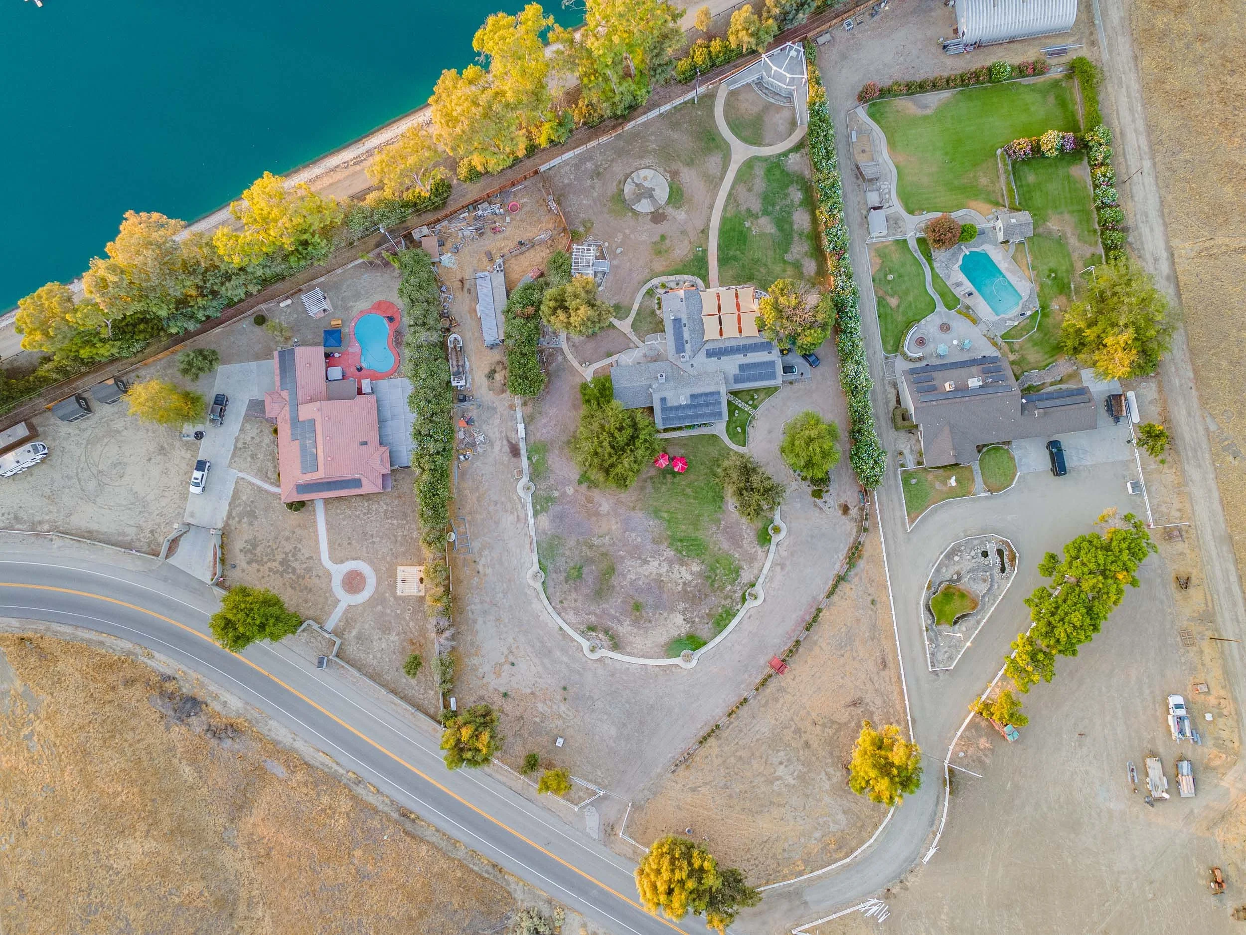Aerial view of a residential property with multiple houses, swimming pools, green lawns, and landscaped gardens, bordered by a body of water and a road.