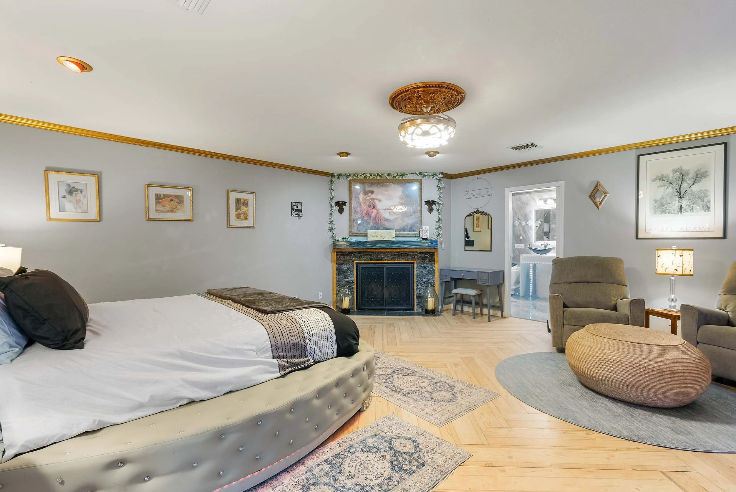 A spacious bedroom with a large bed, beige tufted headboard, black and white bedding, and decorative pillows on the left. The room features hardwood floors, a fireplace with framed artwork above, and various framed pictures and mirrors on the walls. 