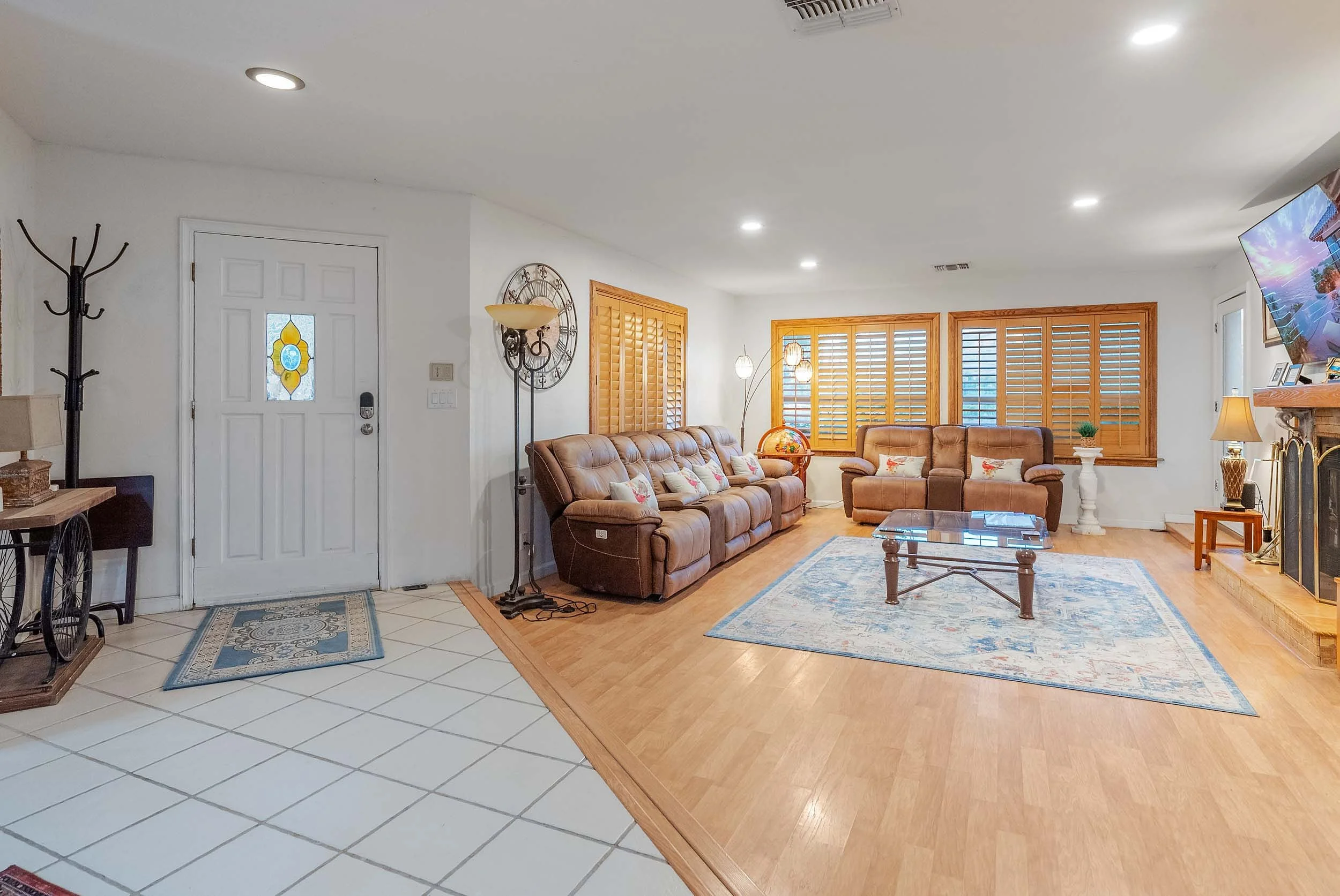 Interior of a living room with brown leather couches, wooden window shutters, a fireplace, area rug, wooden coffee table, decorative items, and a wall-mounted television.