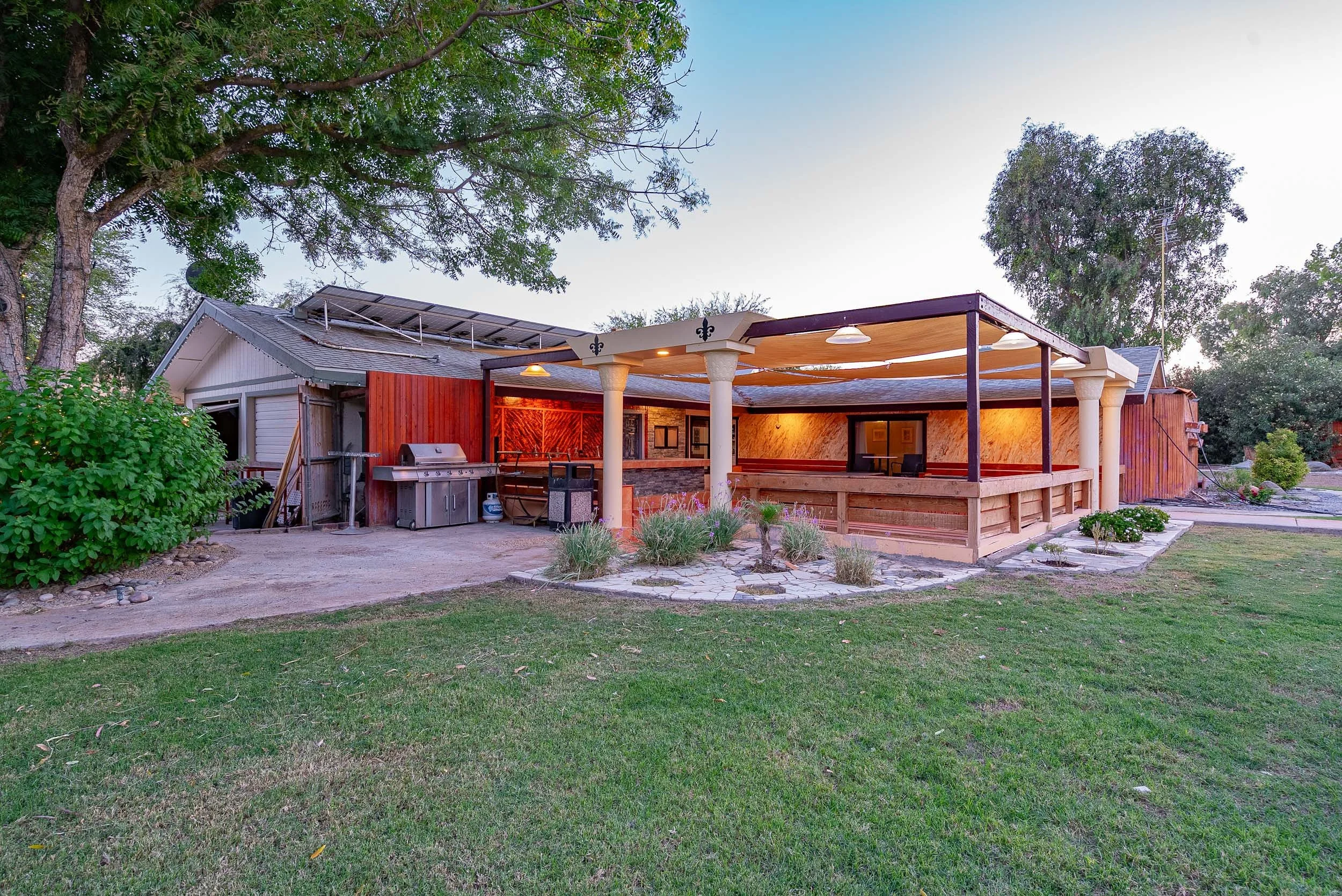 Backyard patio area with a wooden pergola, outdoor grill, and string lights, surrounded by trees and grass.