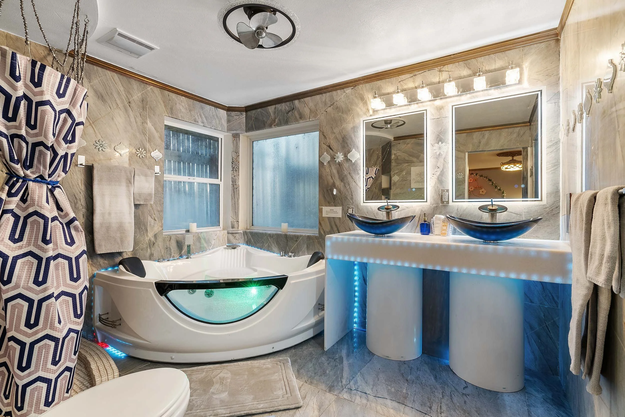 A modern bathroom with marble walls, a bathtub with a curved glass panel, two black vessel sinks on a white illuminated countertop, a large mirror with backlit lighting, and a patterned shower curtain to the left. Two windows provide natural light.