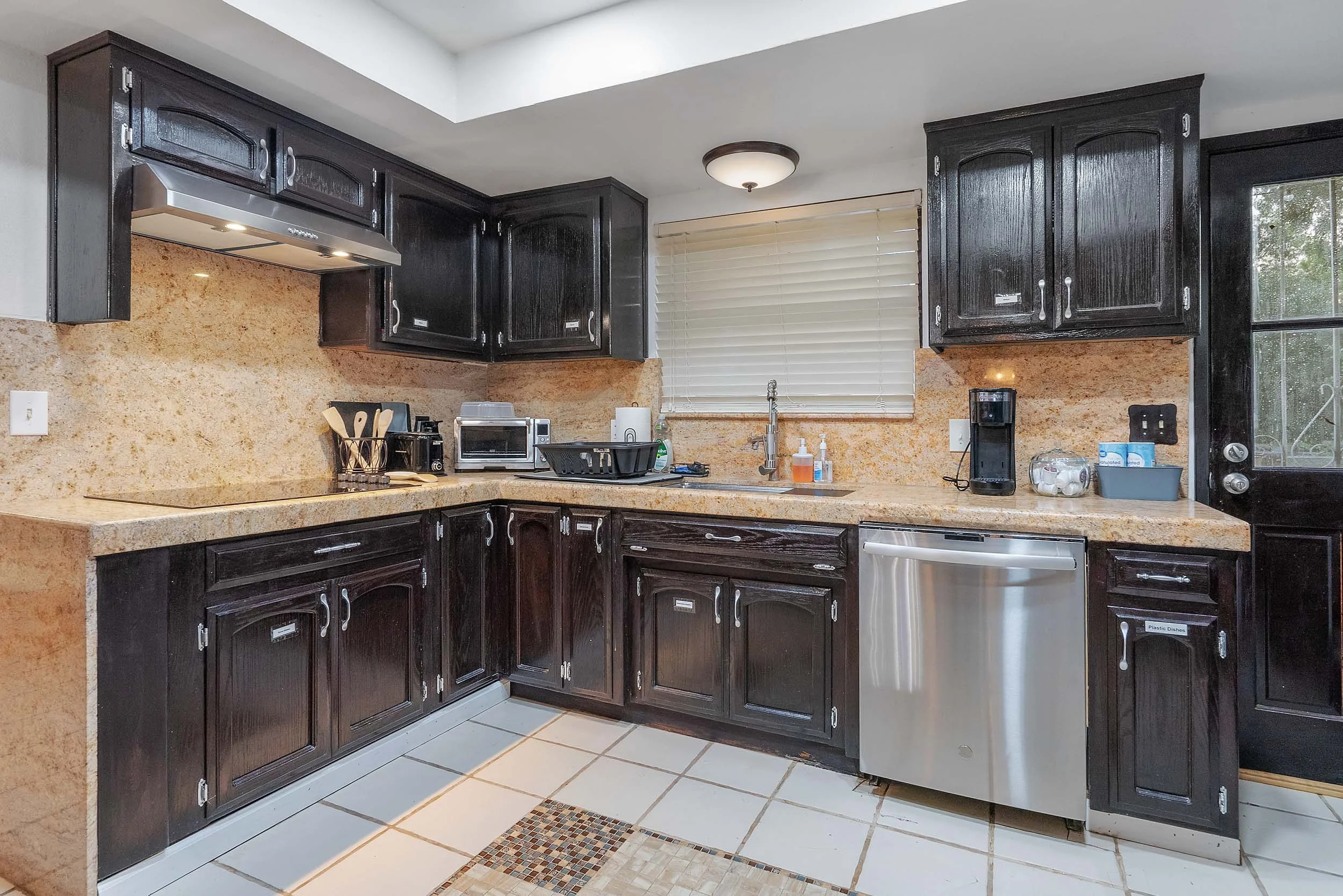 Kitchen with dark wooden cabinets, beige granite countertops, stainless steel dishwasher, black coffee maker, toaster oven, and various small appliances, window with blinds, and tiled floor.