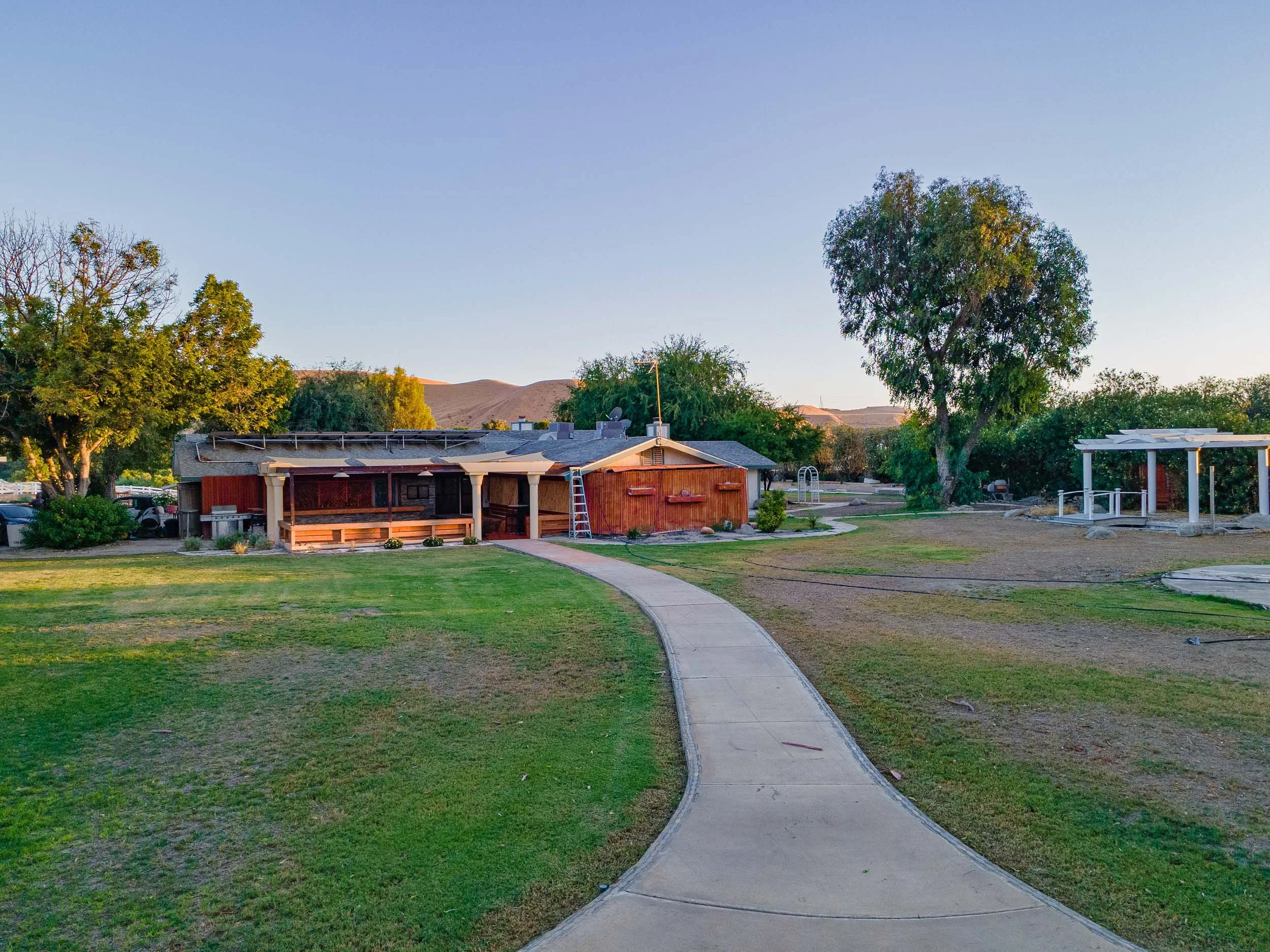 A backyard with a concrete walking path leading towards a house, surrounded by trees and grass, with some structures and hills in the background.