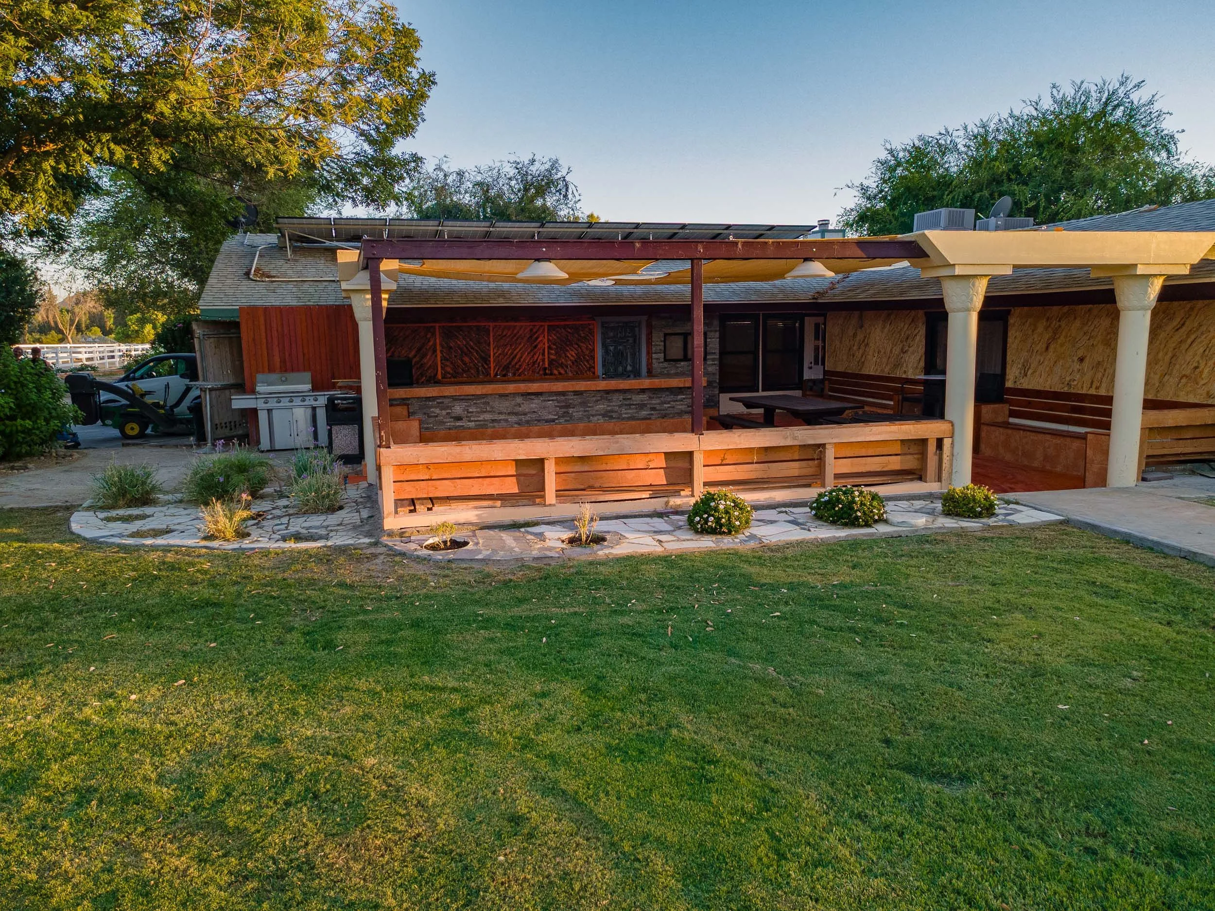 Backyard with patio area, garden plants, and lawn, with an outdoor grill and seating area covered by a roof, in a residential neighborhood.