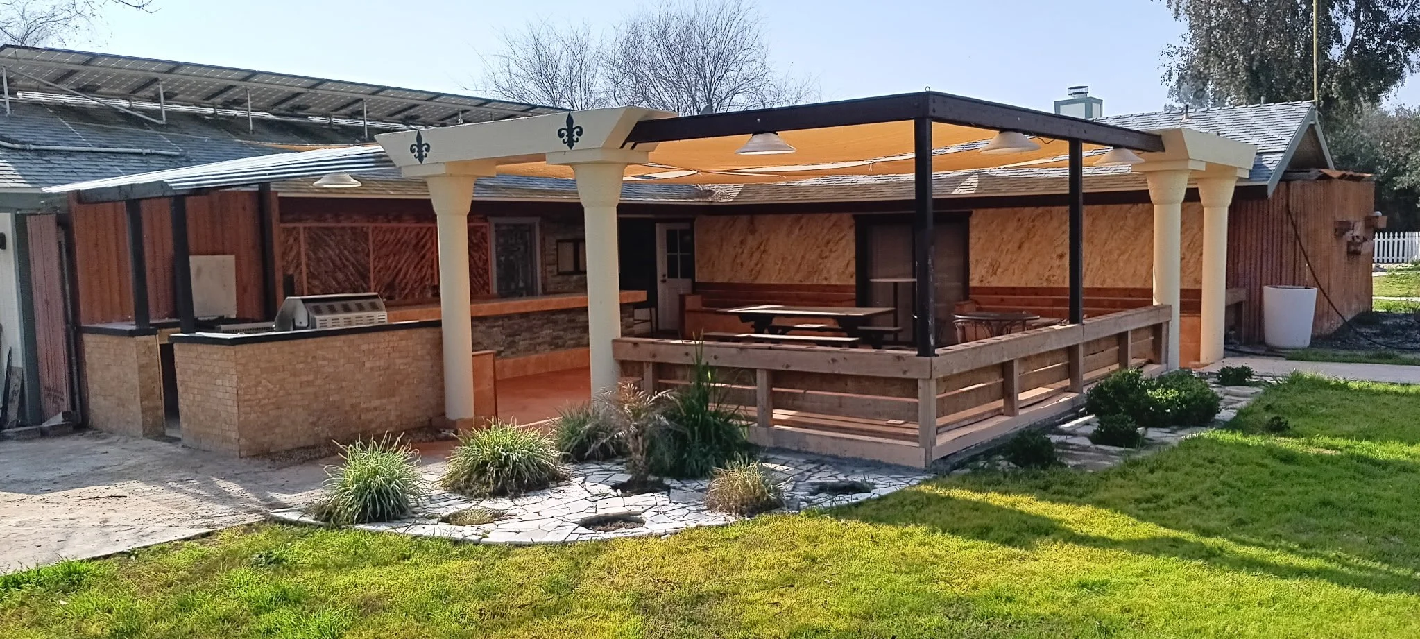 Backyard with patio area, garden plants, and lawn, with an outdoor grill and seating area covered by a roof, in a residential neighborhood.