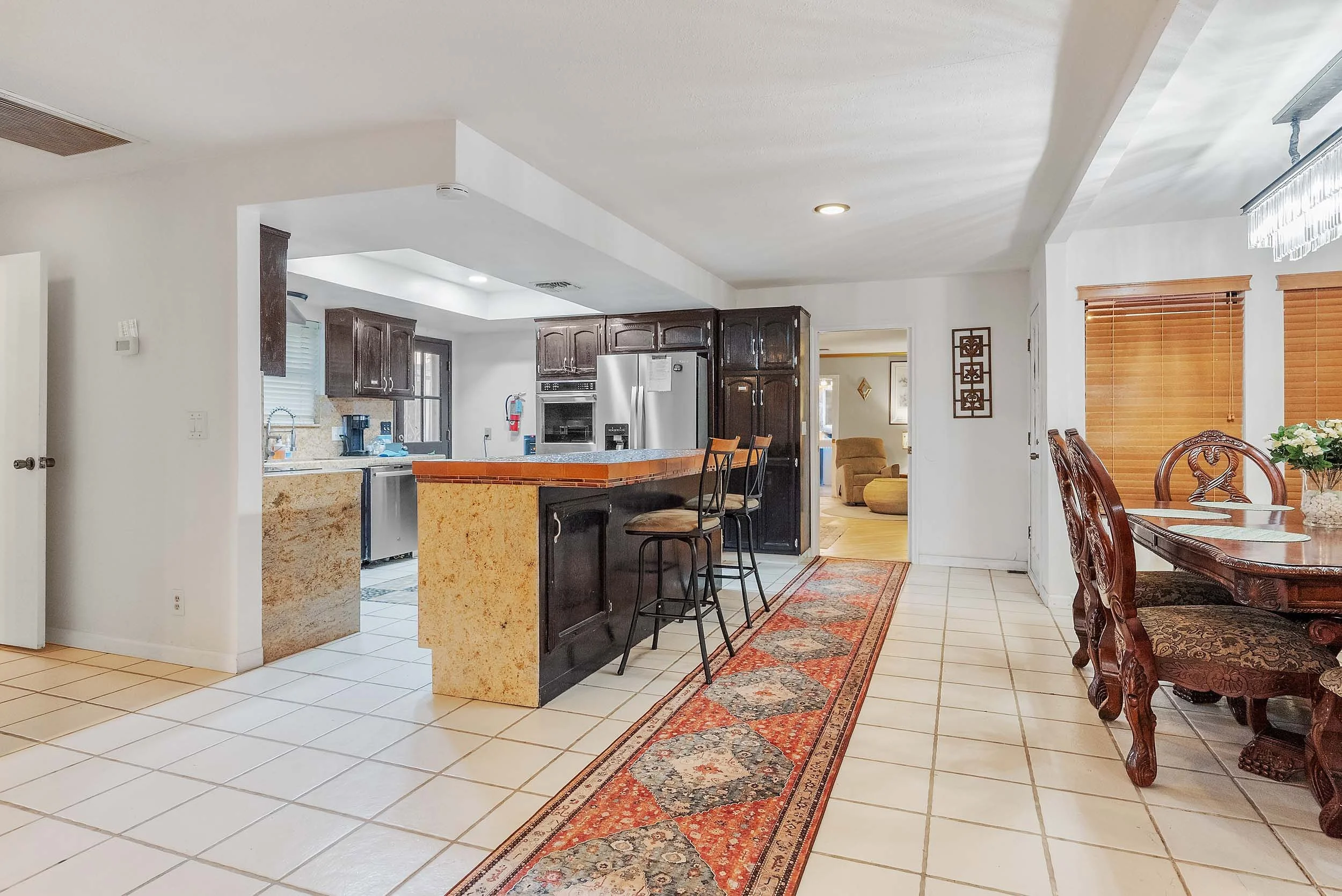 Interior view of a kitchen and dining area with white tile flooring, dark wooden cabinets, stainless steel refrigerator, and a granite kitchen island with two bar stools. To the right is a wooden dining table with ornate chairs and a window with wood