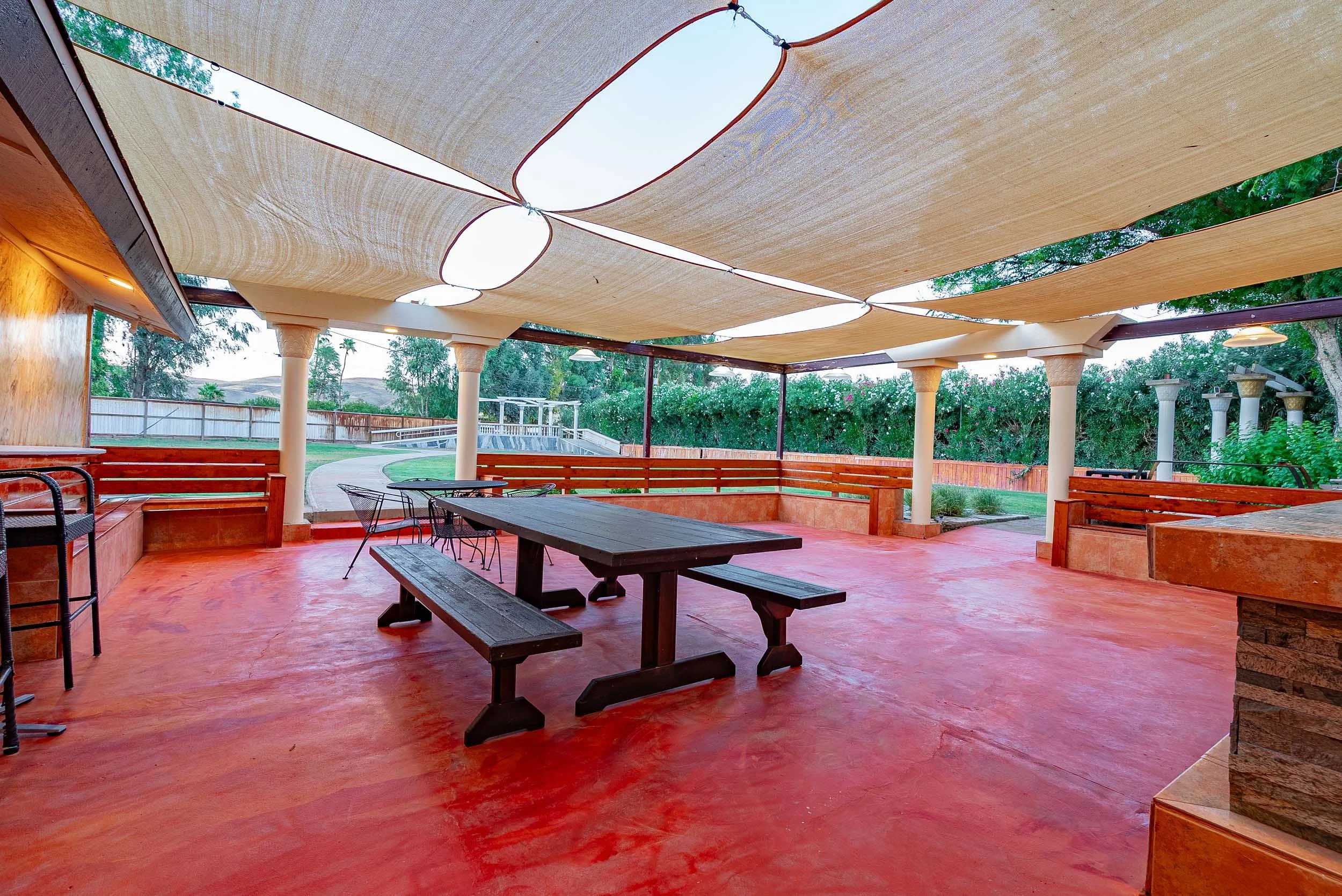 Covered outdoor patio area with wooden benches, tables, and chairs, surrounded by greenery, with a concrete pathway in the background.
