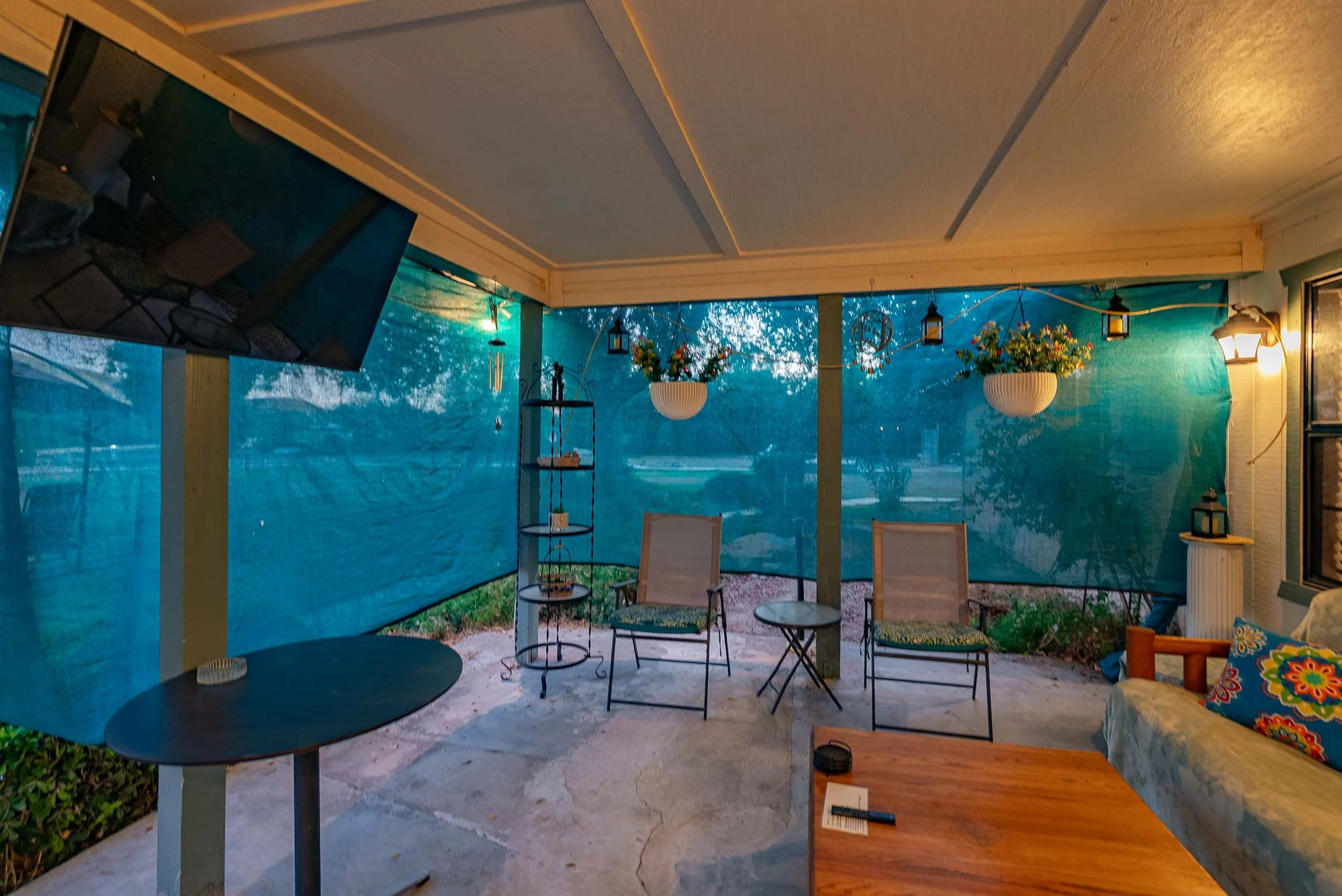 Covered patio with hanging flower baskets, outdoor lanterns, two chairs, a small round table, a couch with a colorful pillow, a security camera, and a large TV mounted on a support pole; blue privacy screens surround the area.