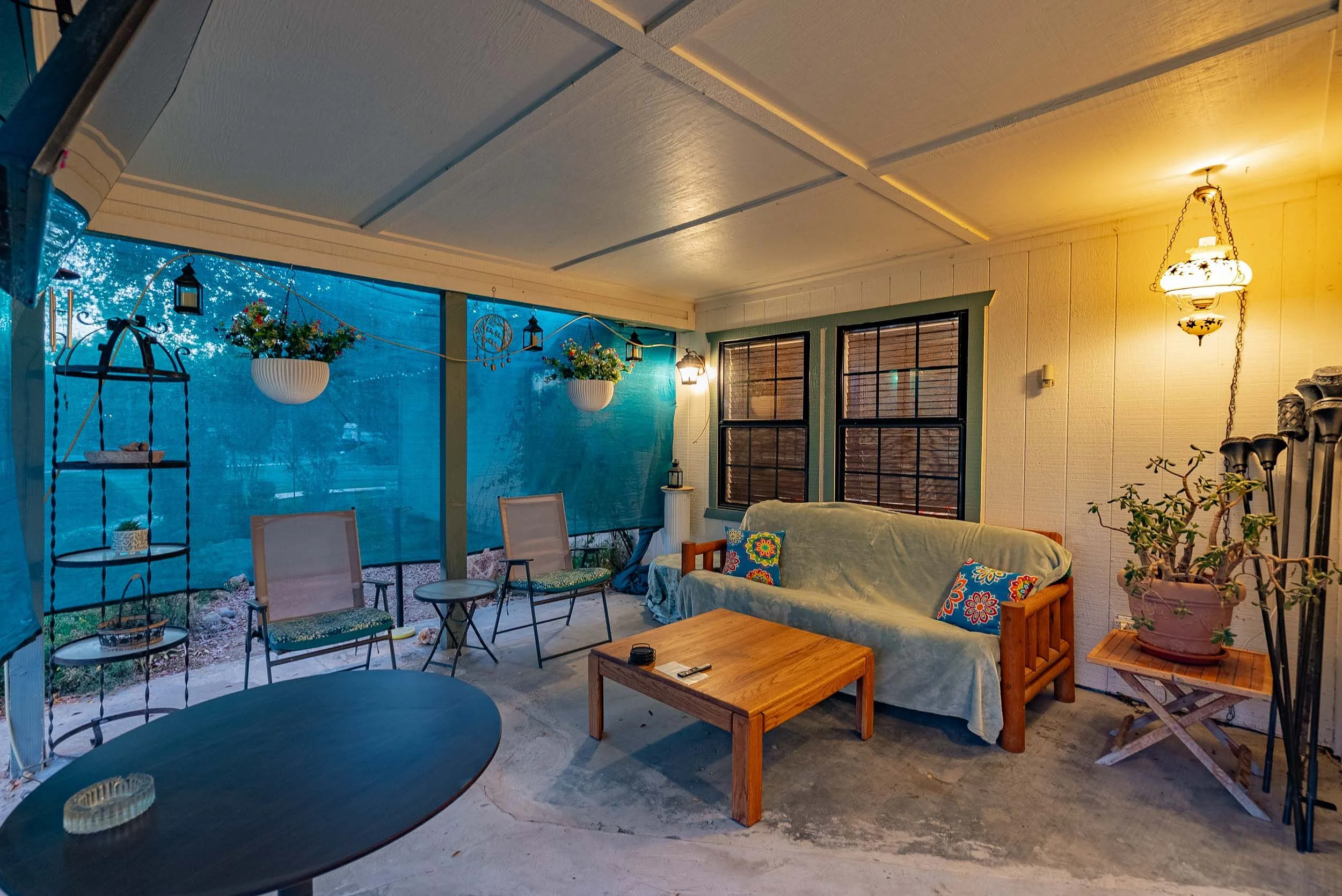 Enclosed patio with outdoor furniture, hanging flower pots, chairs, a couch with colorful pillows, a small wooden table, and warm lighting.