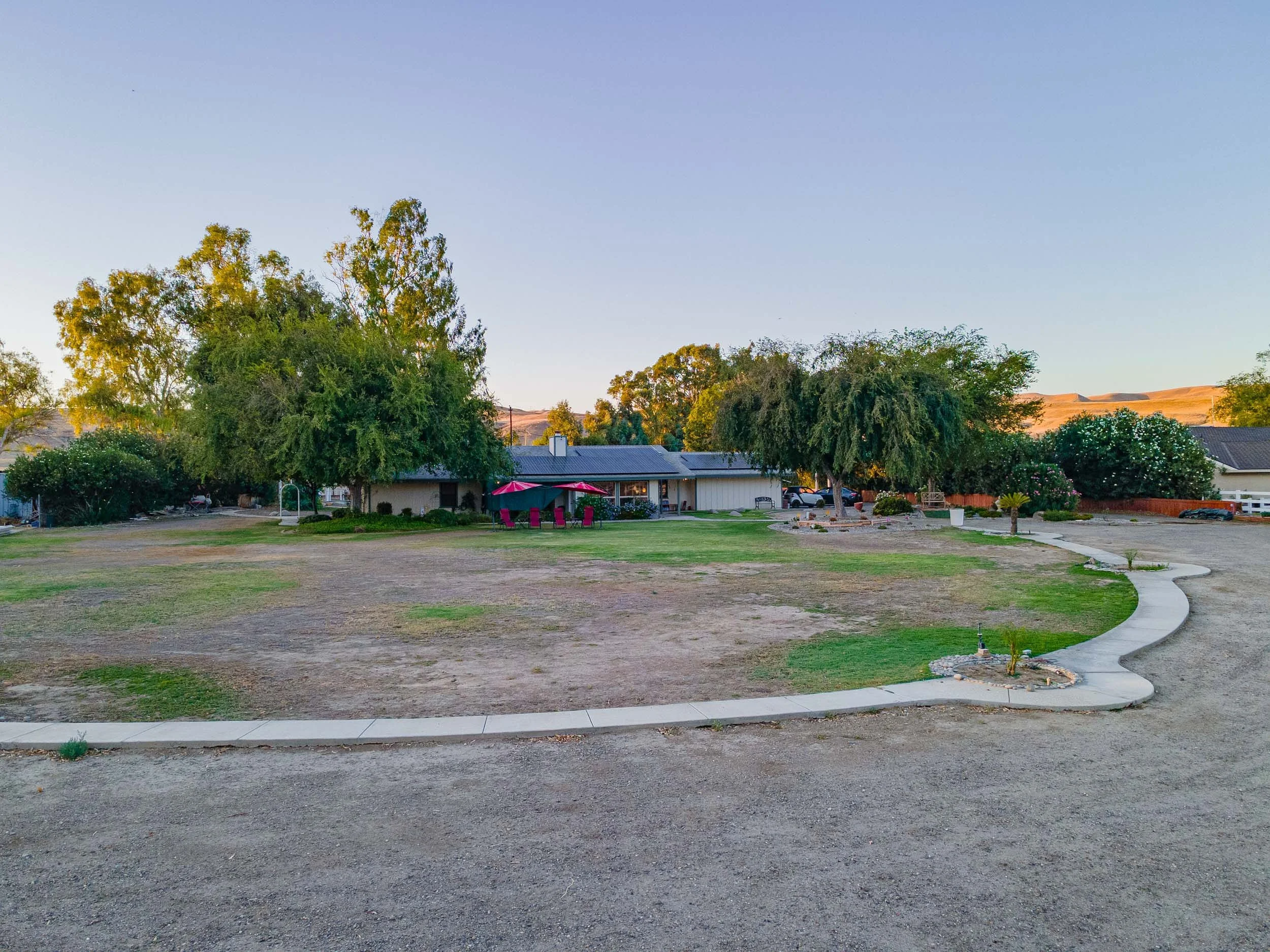 A residential yard with trees, a house with outdoor seating, and a curved sidewalk. The lawn has patches of grass and dirt, with a gravel driveway on the right.