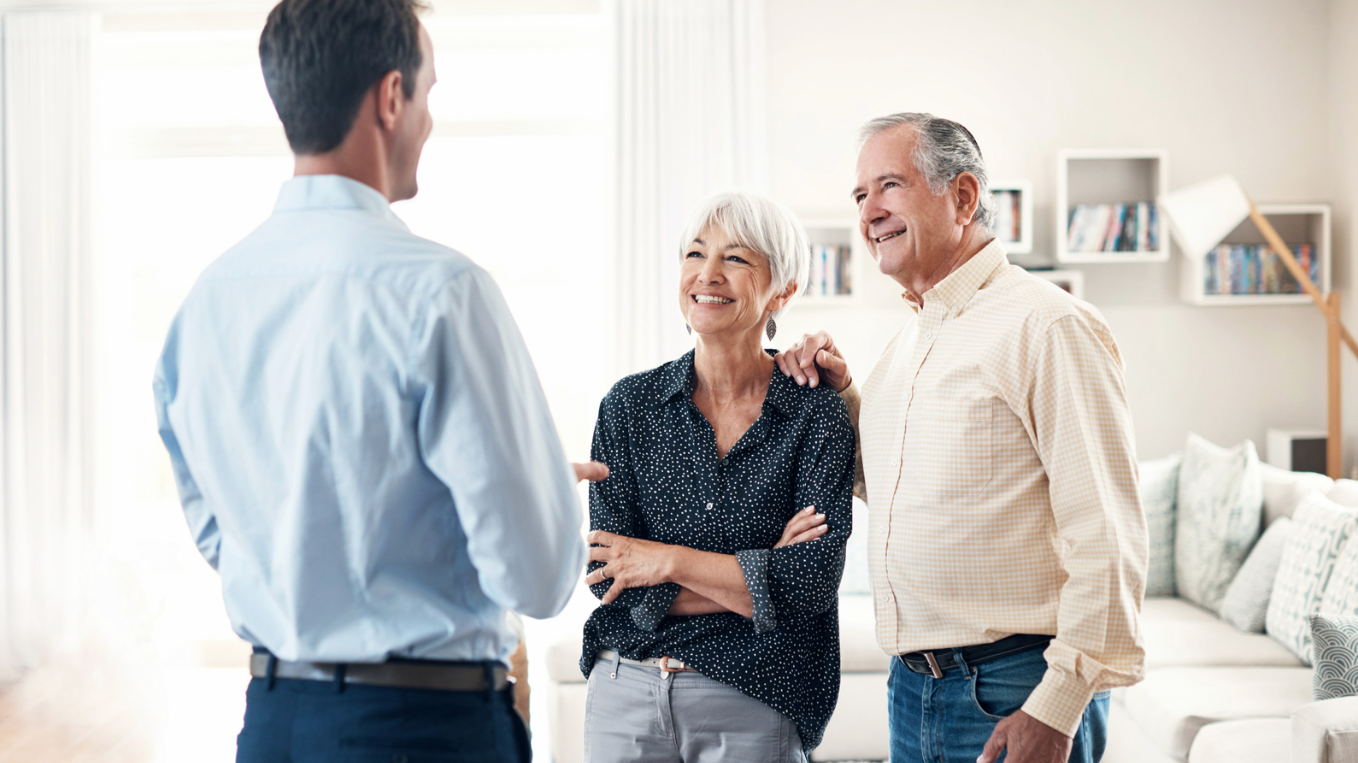 A young man talking to an elderly couple in a living room with sunlight, white walls, and bookshelf.