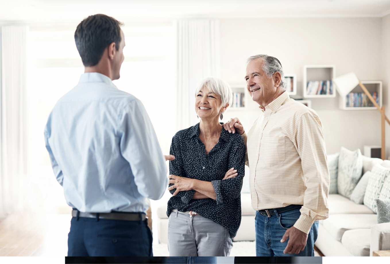An elderly couple talking with a young man indoors, all smiling, bright living room with shelves and a sofa in the background.