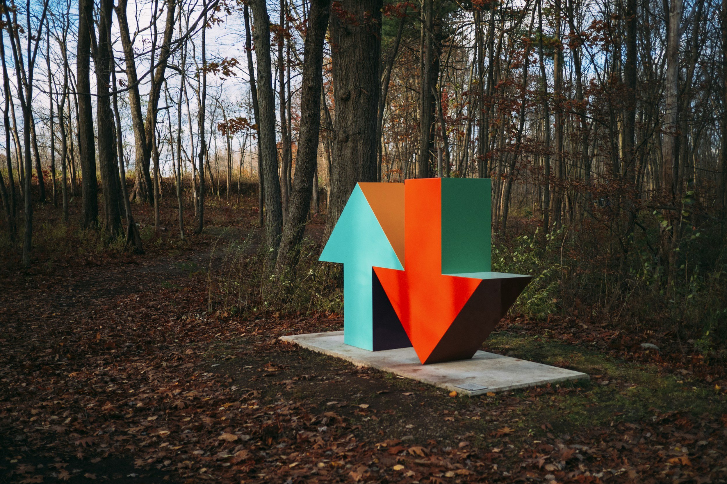 Colorful geometric sculpture with two arrows, one pointing up and the other down, placed in a forest with leaf-covered ground and leafless trees.