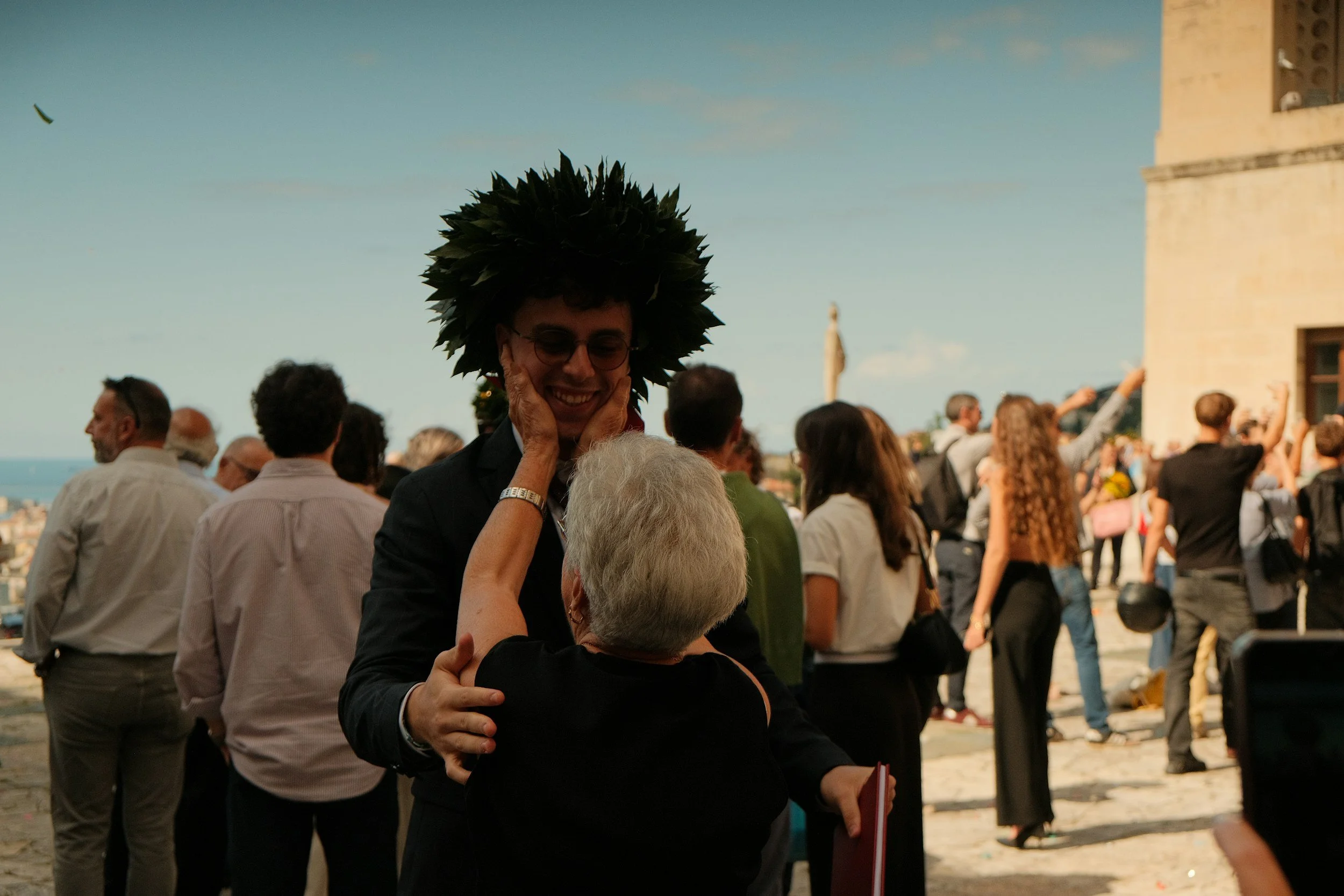 A young man with glasses and a leafy wreath on his head is smiling as an older woman touches his face. They are surrounded by a crowd of people outdoors near a historic building on a sunny day.