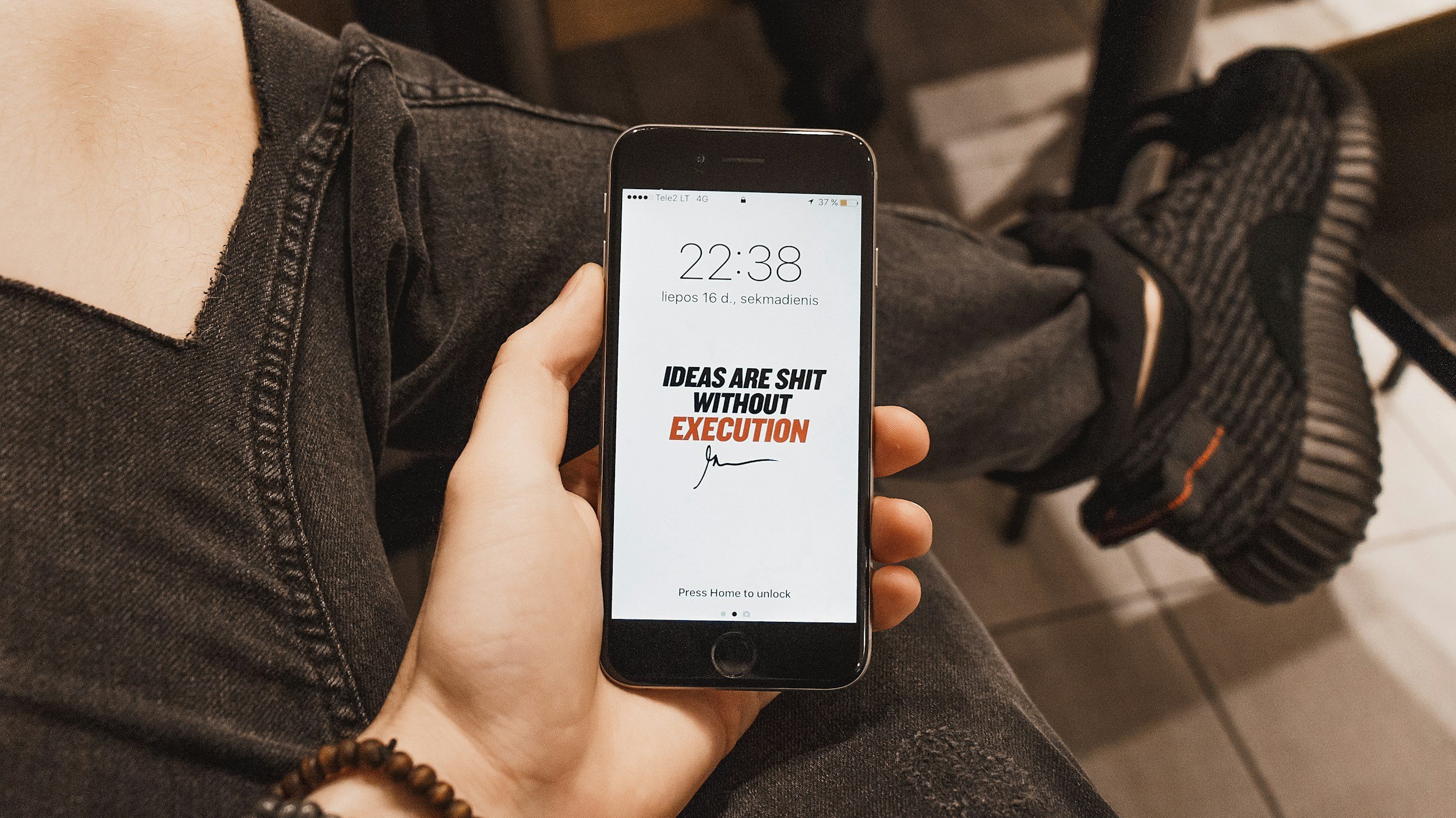 A person sitting and holding a smartphone with a motivational quote on the screen that says, "Ideas are shit without execution," on a digital lock screen at 10:38 PM. The person is wearing black or dark gray ripped jeans and a bracelet, with a backpack on the floor nearby.