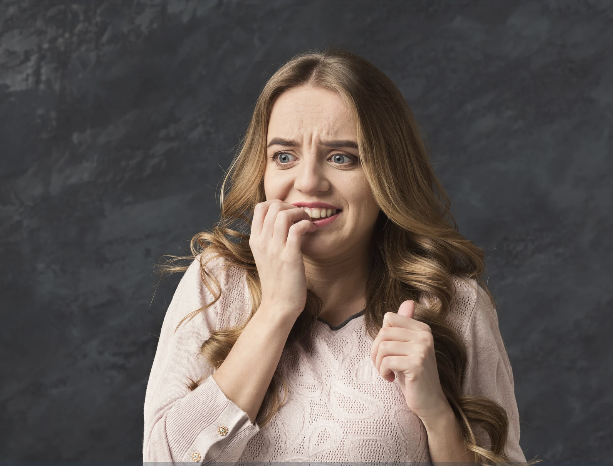 A young woman with long, wavy, light brown hair, wearing a light pink lace sweater, looks worried and anxious, biting her nails and clutching her fist against her chest, against a dark textured background.