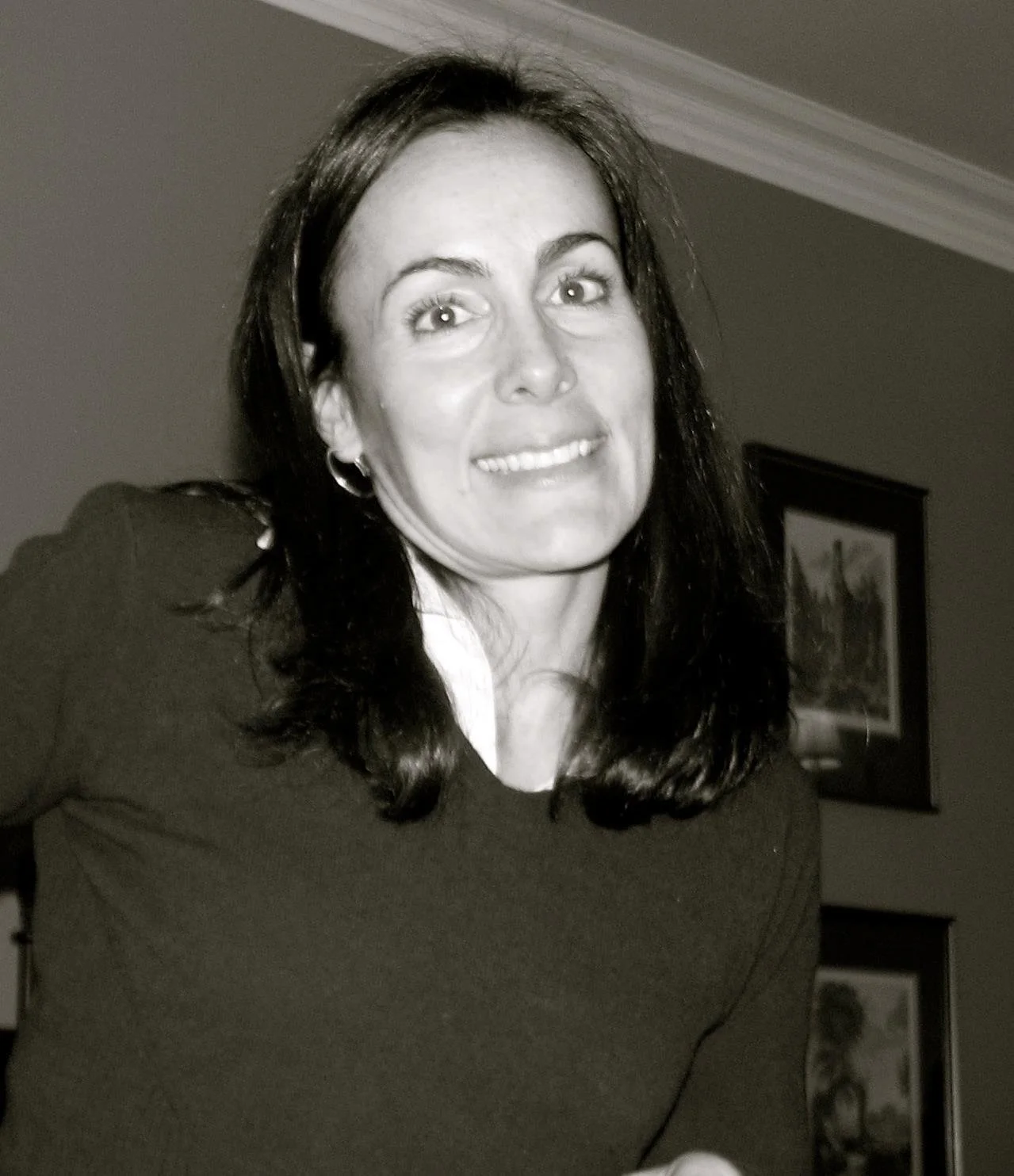 Black and white photo of a woman with shoulder-length dark hair, smiling, wearing a dark top, indoors with framed pictures on the wall behind her.