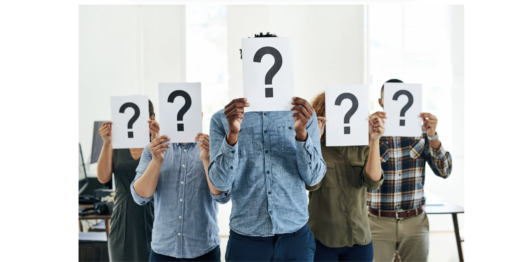 People standing in a row holding signs with large question marks, covering their faces, inside a bright room.