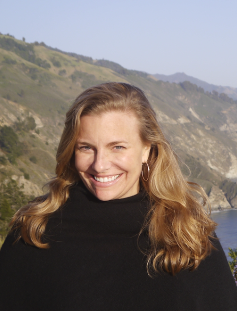 A woman with long blonde hair smiling outdoors with a hillside and water in the background.