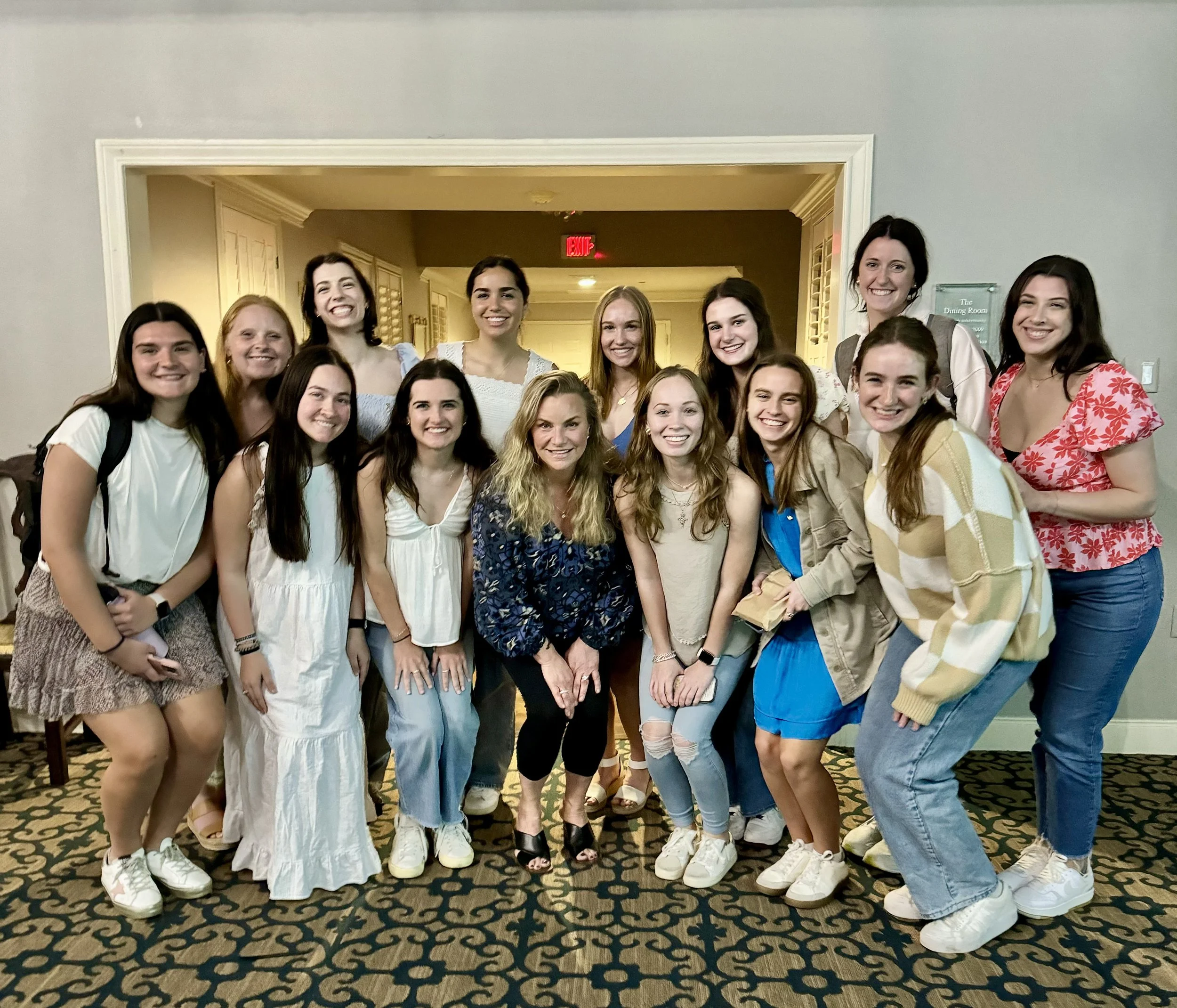 Group of 15 women smiling for a group photo indoors, standing on a patterned carpet near a doorway with a red exit sign in the background.