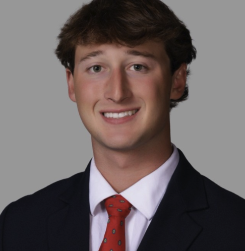 A young man in a business suit with a white shirt and red tie, smiling against a gray background.