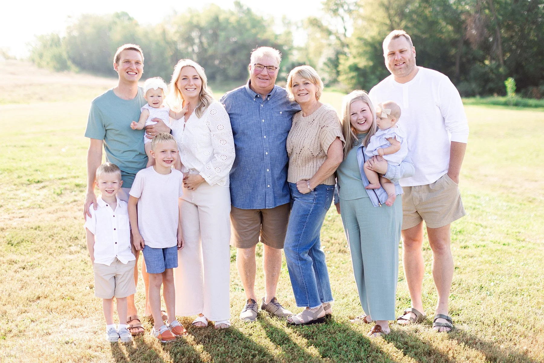 Family gathered outdoors on a sunny day, smiling. The group includes three generations with children, parents, and grandparents, standing on grass with trees in the background.