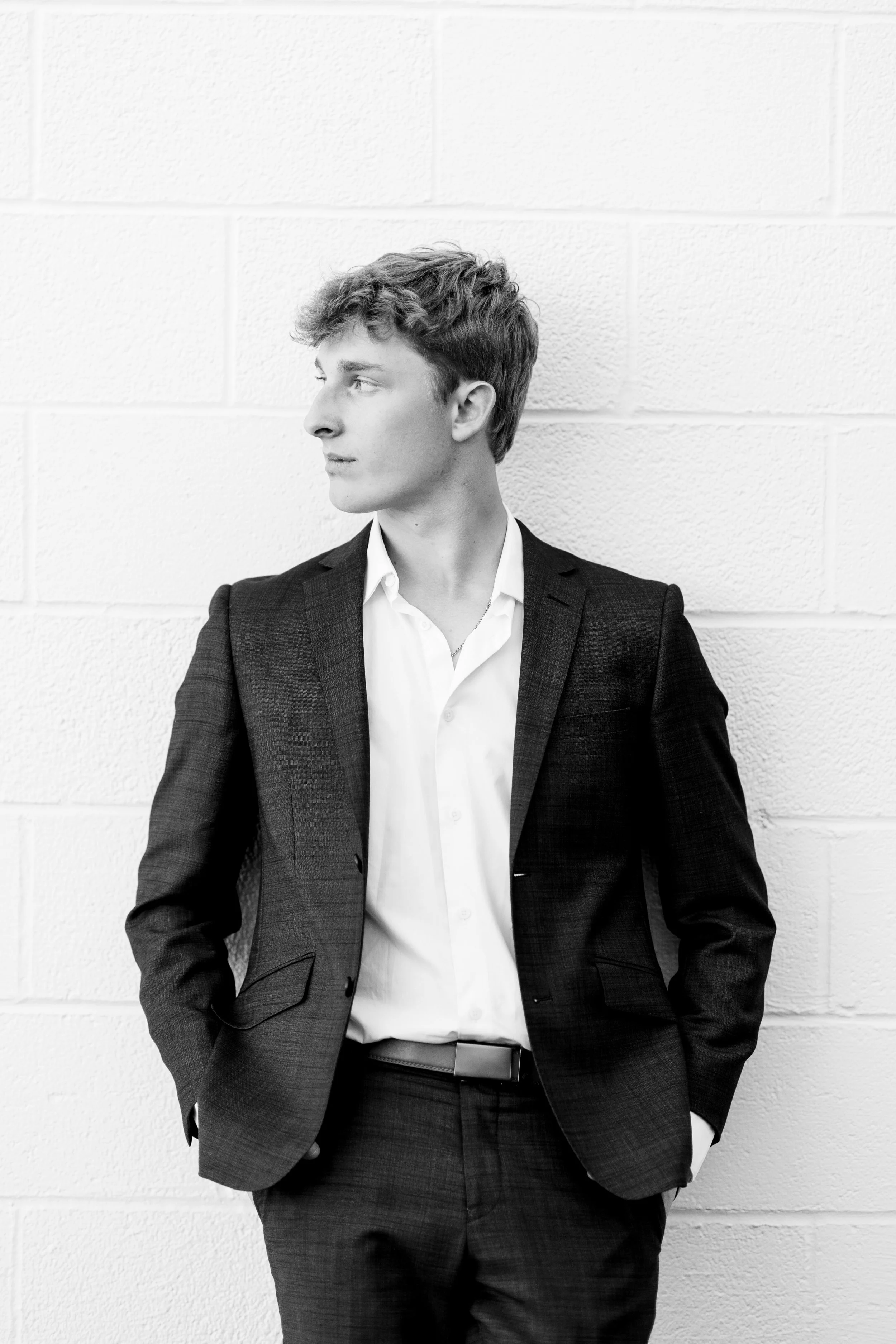 A young man with curly hair wearing a dark suit and white shirt, standing against a white brick wall, looking to his left with hands in his pockets.