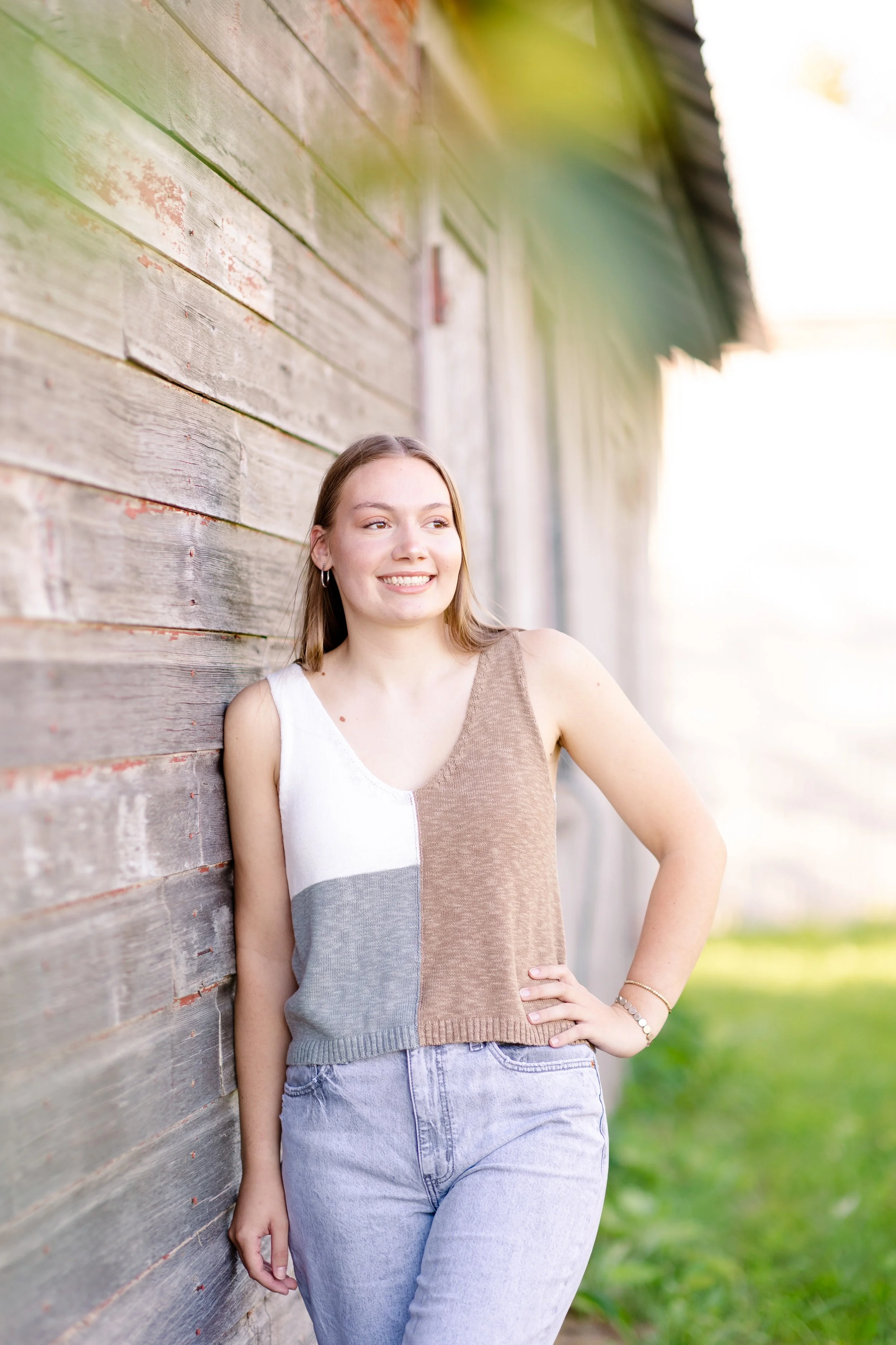 A young woman with long hair and hoop earrings leaning against a weathered wooden wall outdoors, smiling and looking to the side.