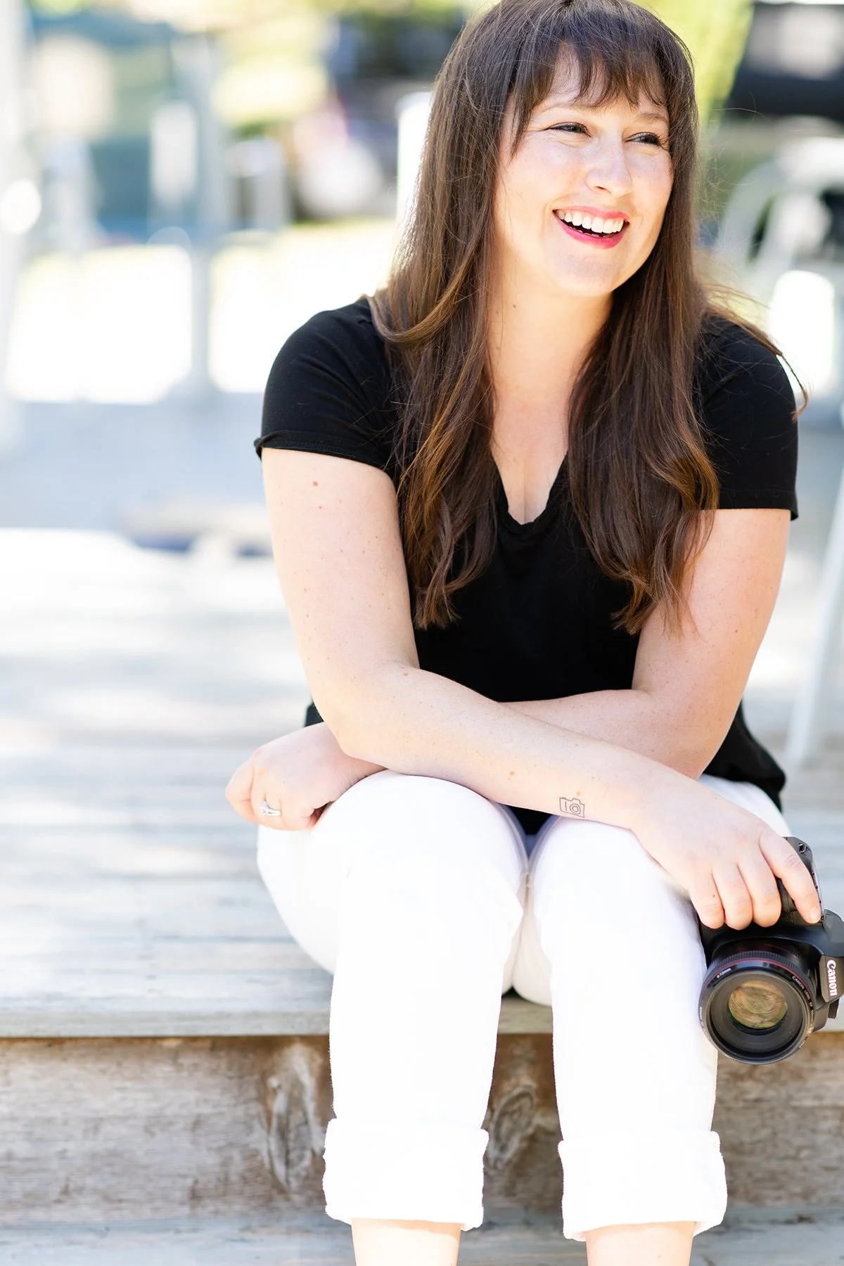 A woman with long brown hair, wearing a black shirt and white pants, sitting on a wooden dock outdoors, smiling and holding a professional camera with a telephoto lens.