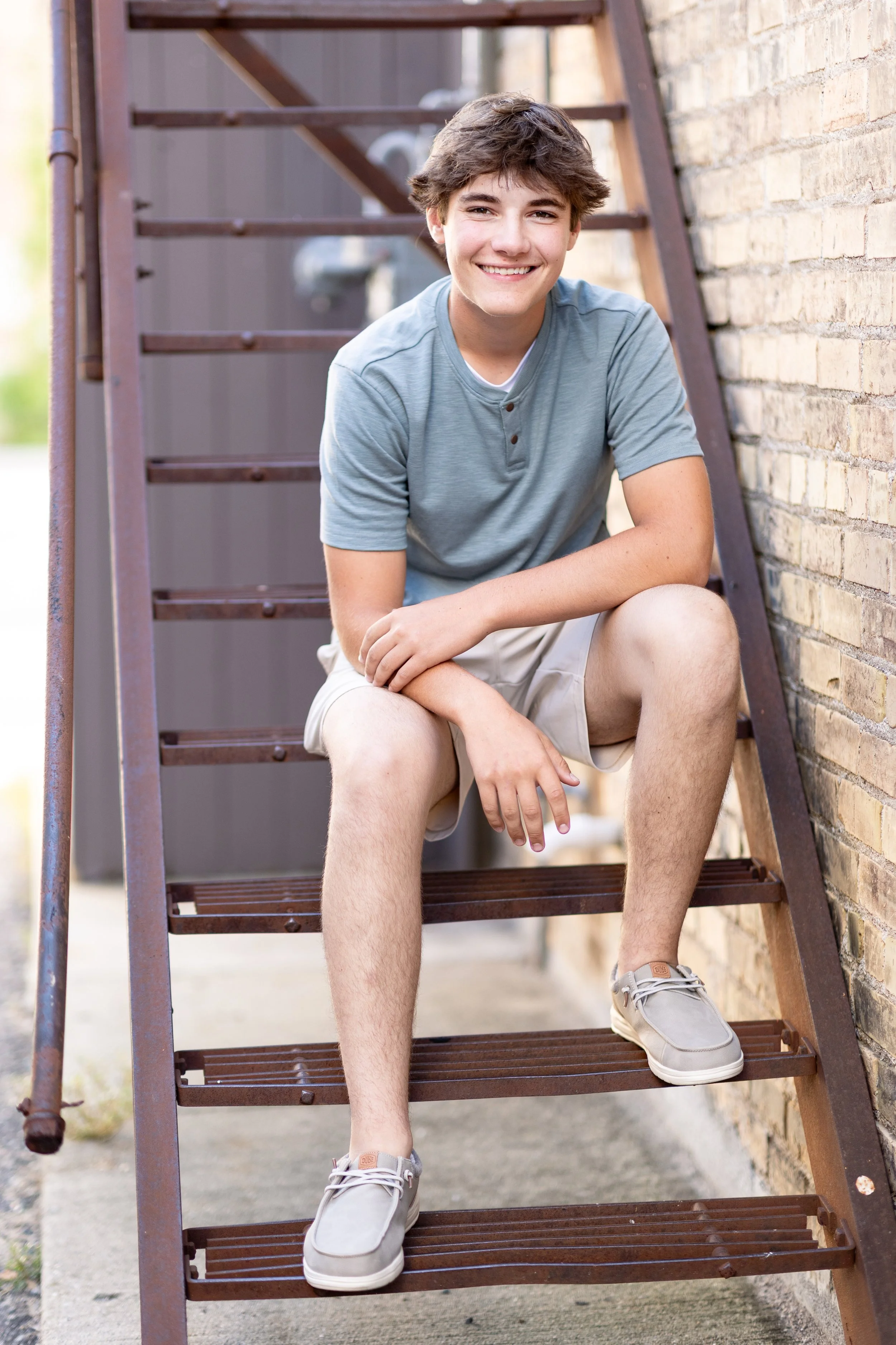 A teenage boy sitting on outdoor metal stairs next to a brick wall, smiling, wearing a light blue t-shirt, beige shorts, and gray sneakers.