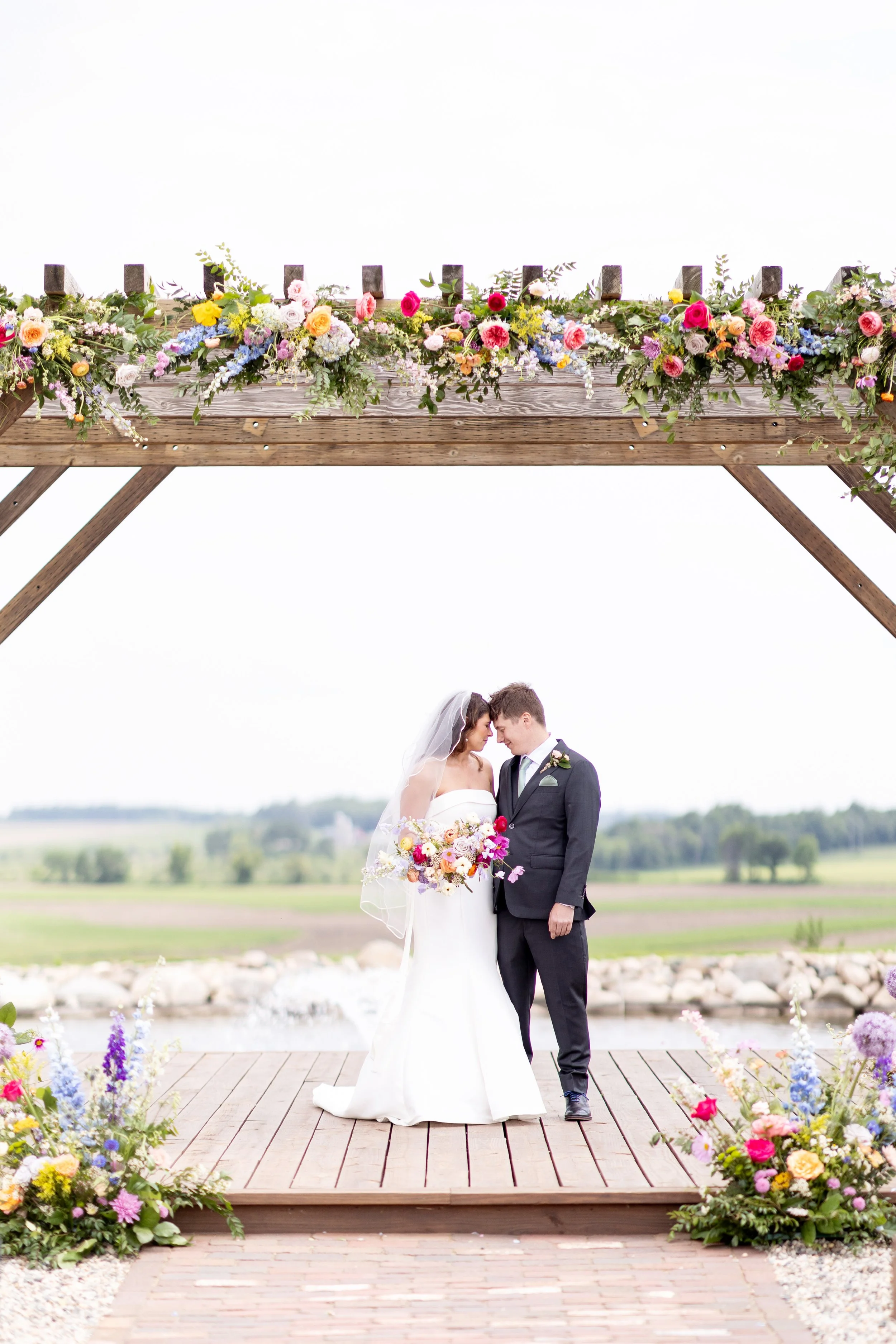 A bride and groom stand close together on a wooden deck overlooking a field, sharing a moment beneath a floral arch. The bride is wearing a white strapless wedding gown and veil, holding a colorful bouquet, while the groom is dressed in a dark suit with a boutonniere. The arch above them is decorated with a variety of colorful flowers and greenery.