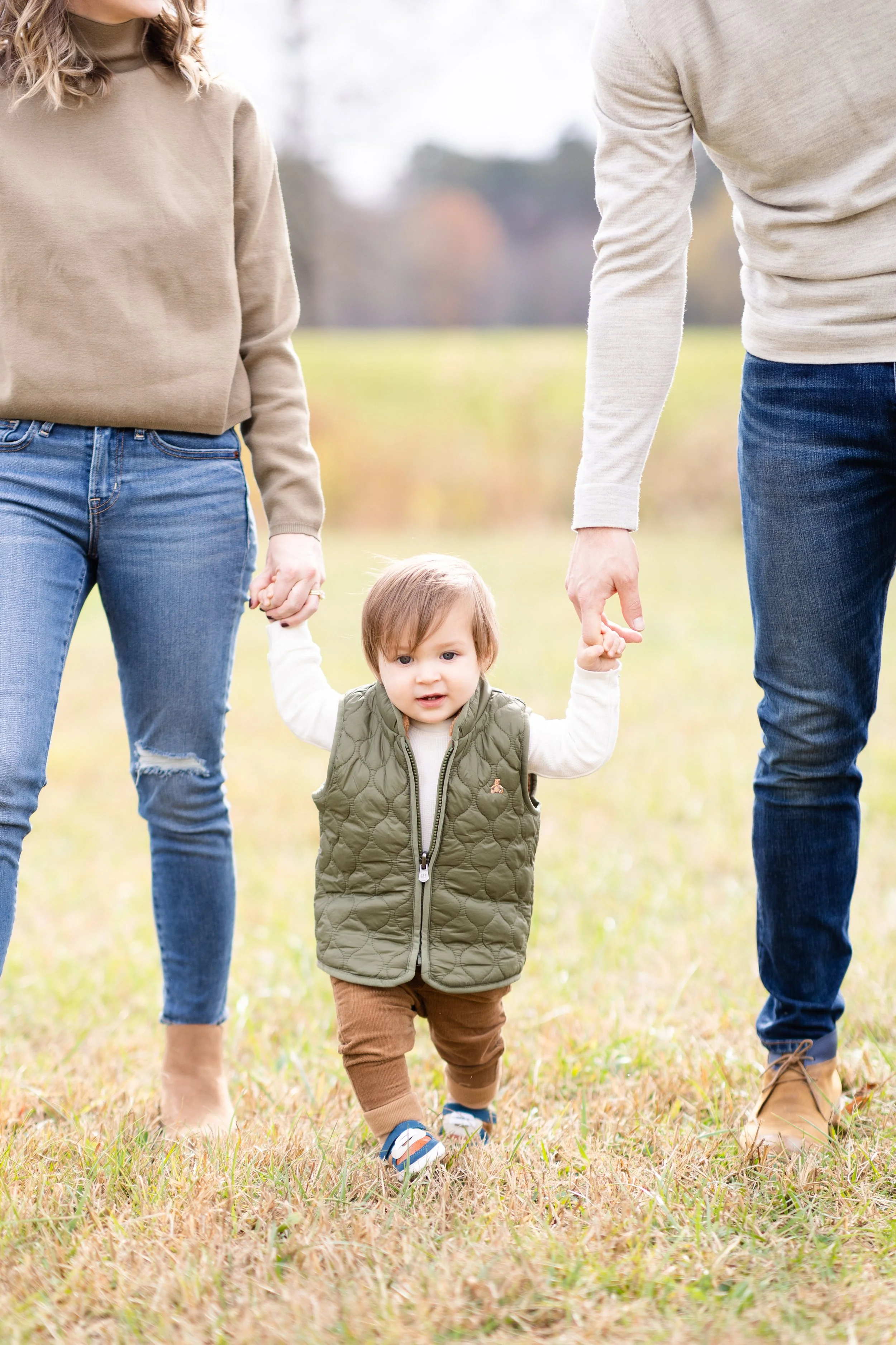 A young child is walking outdoors on grass while holding hands with two adults, one on each side, in an open field with blurred trees in the background.